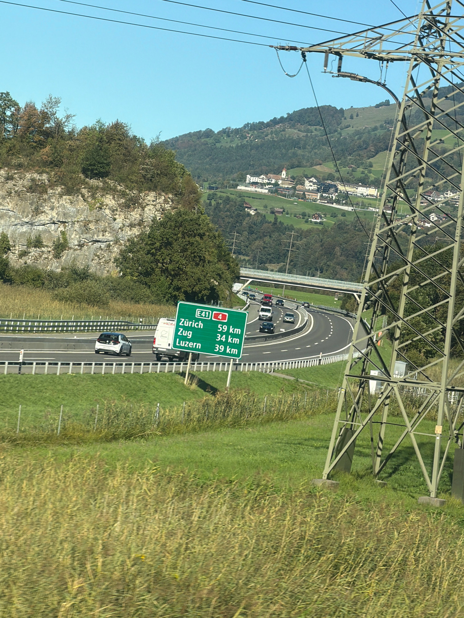 The image depicts a view of a highway in Schwyz, Switzerland, with a large electrical pylon dominating the right side of the frame. The highway is curved and has several lanes of traffic, with cars and a white van visible. A green directional sign indicates distances to Zürich (59 km), Zug (34 km), and Luzern (39 km). In the background, rolling green hills are dotted with houses and a church, suggesting a rural or semi-rural setting. The sky is clear and blue, and the overall lighting indicates daytime. The foreground is filled with tall, dry grass.