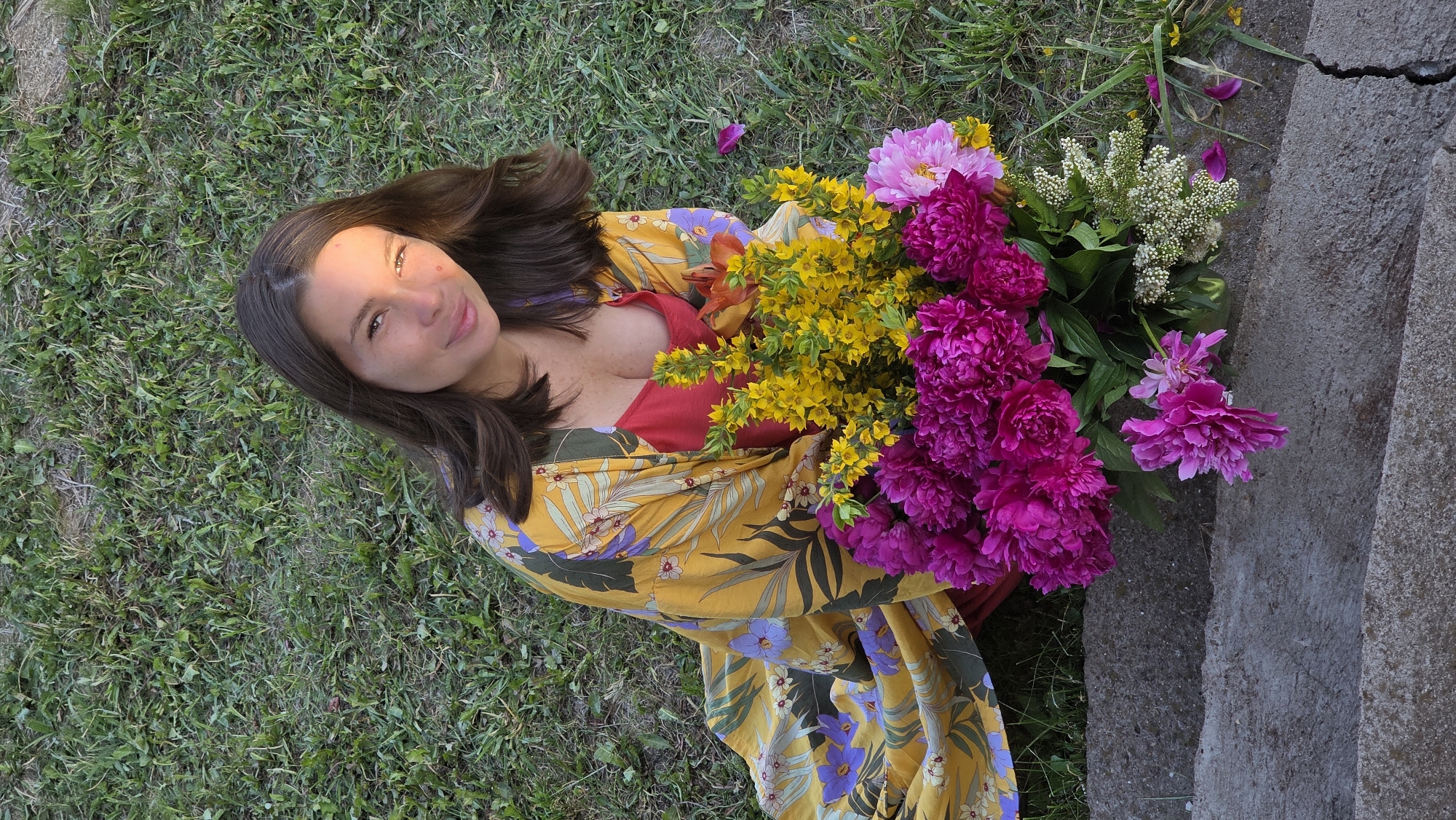 A young woman with long, dark hair lies on a grassy surface, propped up on her elbow. She is wearing a yellow and purple floral robe over a red top. Her face is turned towards the camera, and she is smiling. She holds a large bouquet of vibrant pink and yellow flowers. To her right, a concrete structure, possibly stairs, is visible. The scene appears to be outdoors, with sunlight illuminating the woman's face and casting shadows. The overall mood is serene and picturesque, suggesting a warm, sunny day. The location is identified as Kupoyarovo, Russia. There is no visible text in the image.