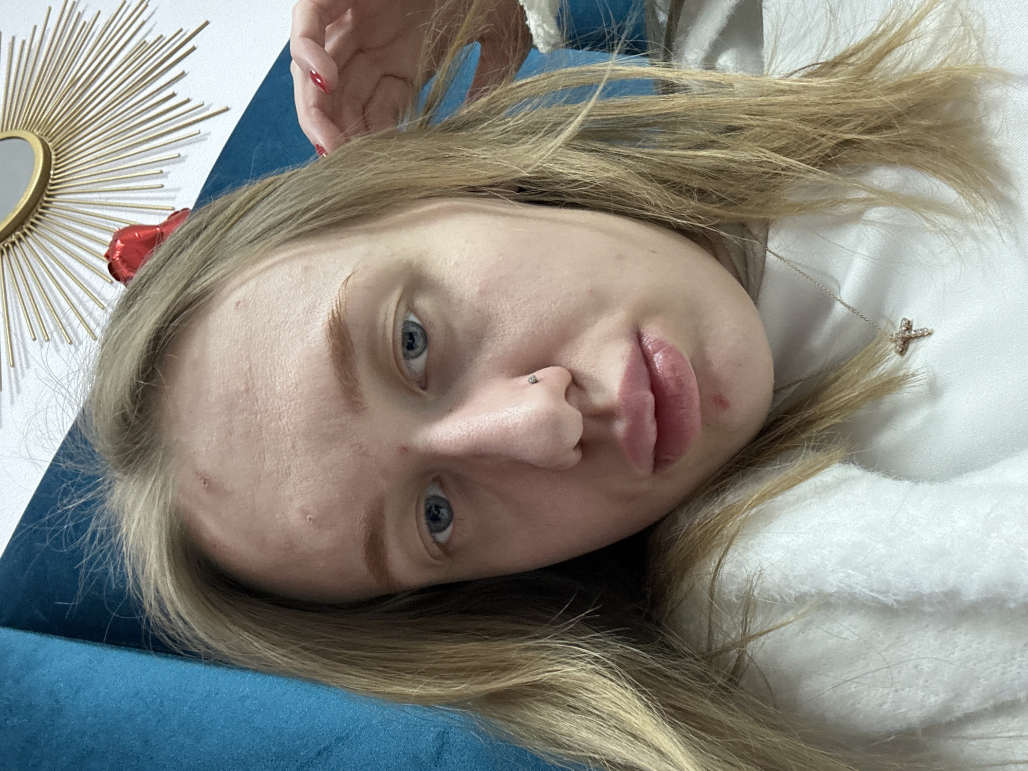 A close-up, angled portrait shows a fair-skinned woman with blonde hair and blue eyes. She is lying down, with her head resting on a blue surface, possibly a cushion or sofa. A portion of a gold sunburst-shaped decoration or mirror is visible in the background. The woman has a nose piercing and wears a delicate necklace with a cross pendant. Red accents, possibly hair accessories or decorations, are visible in her hair. Her expression is neutral, and the focus is on her face and features. There is no indication of the location being Rostov-on-Don, Russia. The scene does not provide enough information to determine the exact location.