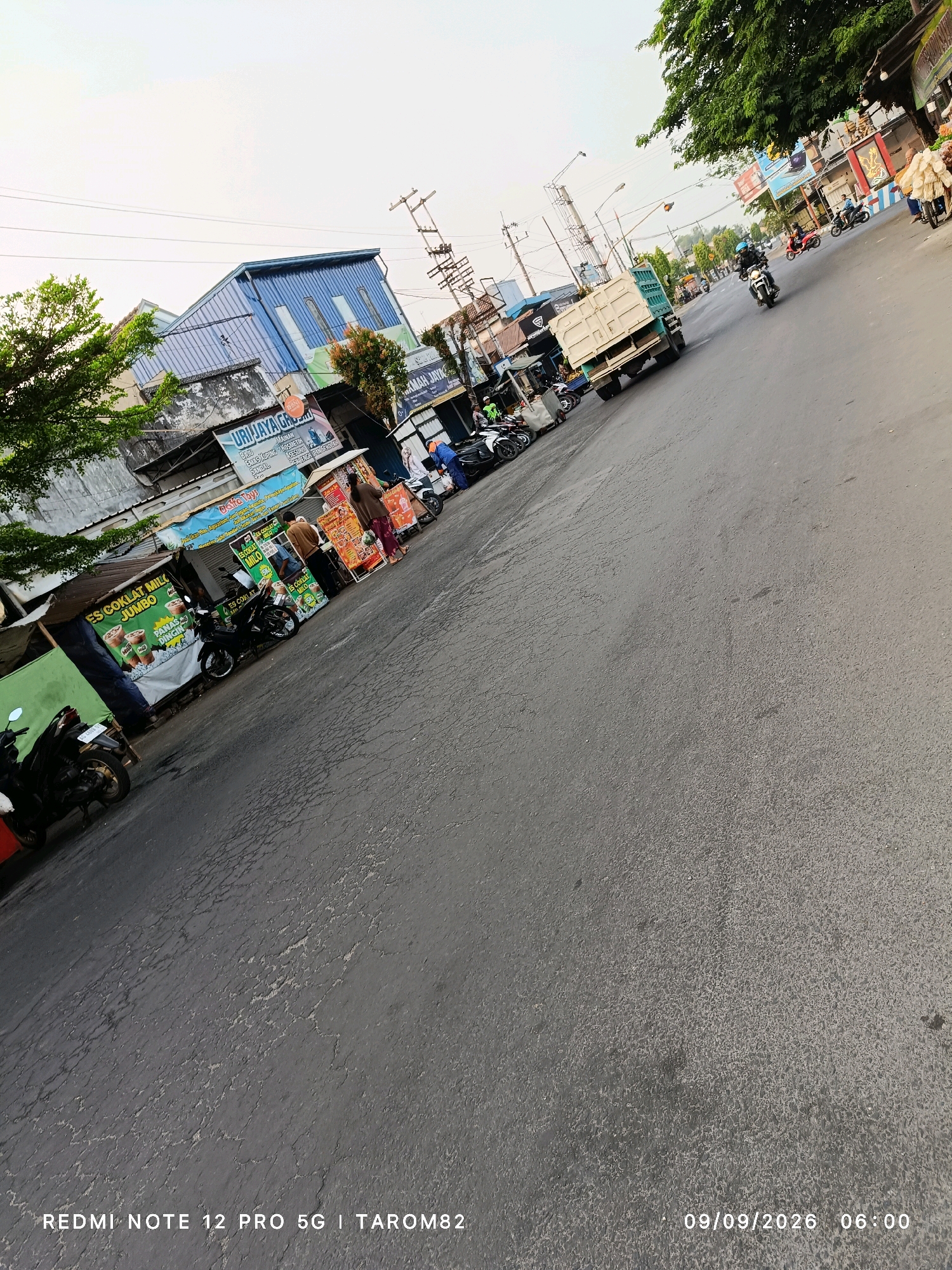 In the city of Ngoro, Indonesia, a street scene unfolds with various elements of daily life. On the left, a row of shops lines the road, displaying advertisements for "ES COKLAT MILO JUMBO" and other food and beverage products. Several motorcycles are parked along the sidewalk, indicating local transport. A man in a blue uniform is visible behind a stall, interacting with customers. Further down the street, a large, light-colored truck is parked, its cargo area empty. Various other vehicles, including motorcycles with riders, are moving along the asphalt road, suggesting a bustling urban environment. The sky is overcast, casting a diffused light on the scene. Overhead, power lines and utility poles are visible, typical of urban infrastructure. The image also captures details of the road surface, which appears worn and cracked in places. The timestamp "09/09/2026 06:00" is embedded in the bottom right corner of the image, along with text identifying the device as "REDMI NOTE 12 PRO 5G | TAROM82".