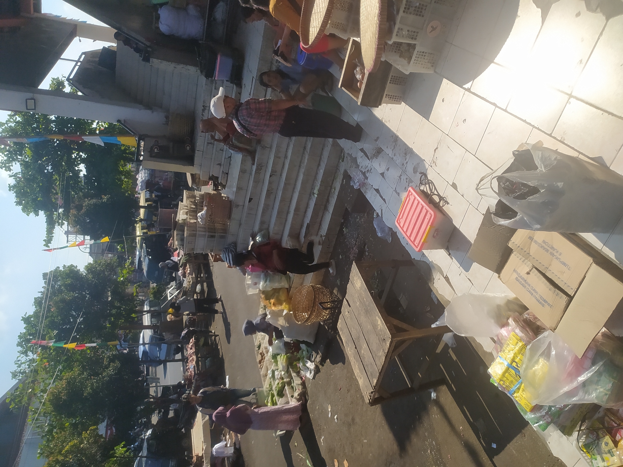 The image depicts a bustling market scene, likely in Kupang, Indonesia, given the presence of flags and typical market structures. In the foreground, a weathered wooden table sits on a tiled ground, casting a sharp shadow. To the left, various goods are displayed, including plastic bags filled with produce and packaged items.
Further into the scene, several individuals are engaged in market activities. A woman in a pink hijab and long dress stands near a stall displaying vegetables in white crates. Another woman, wearing a dark headscarf, sits behind the vegetables. To the right, a person in a red and white plaid shirt and dark pants, wearing a white cap, stands on the steps of a building. Several other people are visible in the background, interacting with stalls or walking through the market.
The environment suggests an outdoor market with some covered areas. The sunlight indicates it is daytime. Flags, possibly representing local or national colors, are strung across the area, adding to the festive or community atmosphere. The overall scene is a snapshot of daily commerce in what appears to be a vibrant Indonesian market.