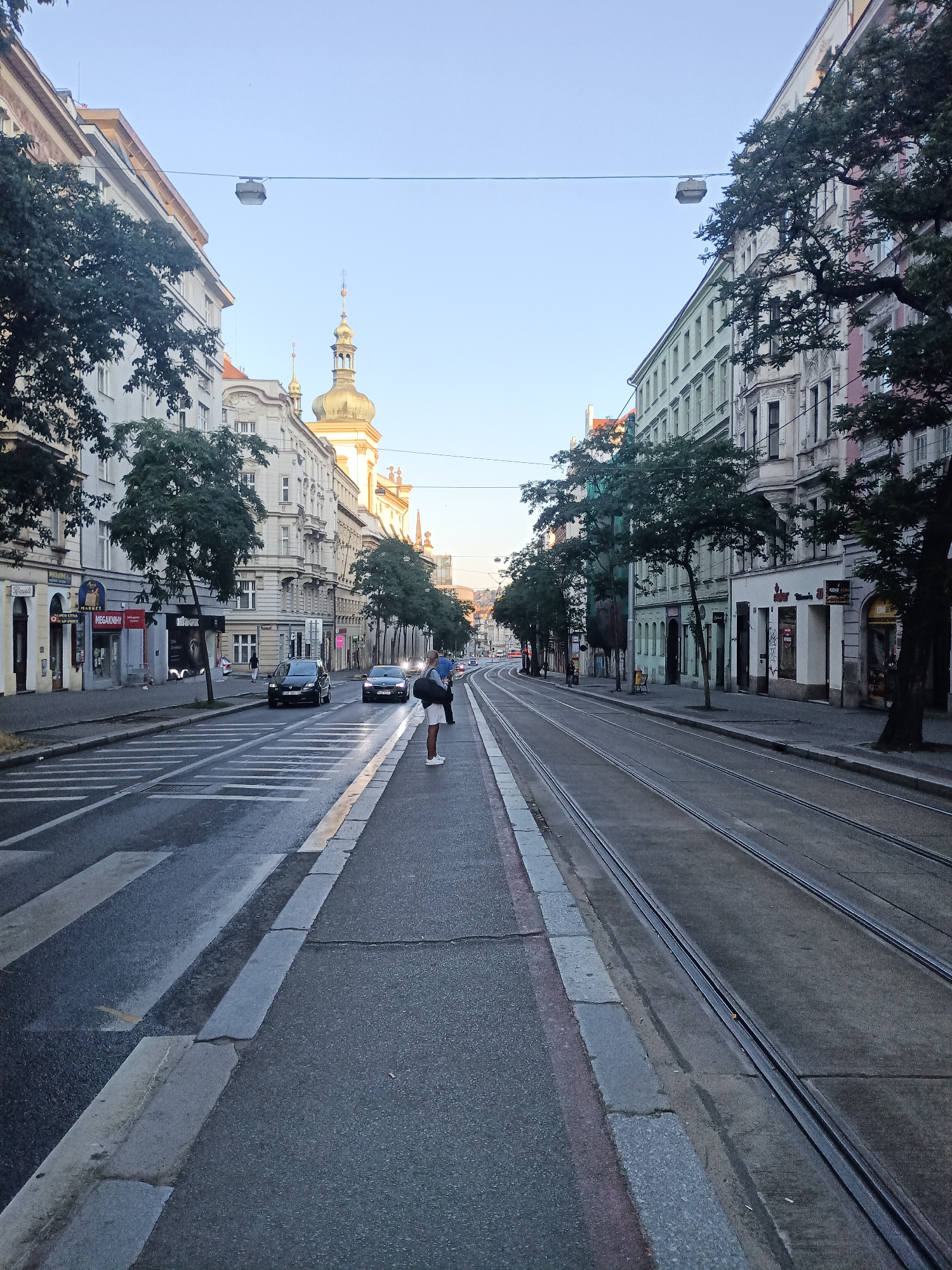 The image captures a view down a city street in Prague, Czechia, characterized by tram tracks running along the right side of the road. The street is lined with multi-story buildings exhibiting traditional European architecture, with ornate facades and upper floors. Trees are planted along the sidewalks, some of which are partially obscured by scaffolding.
In the foreground, a pedestrian is standing on a crosswalk, facing away from the camera. The pedestrian is wearing dark shorts, a white t-shirt, and white sneakers, with a backpack slung over one shoulder. Two cars are visible on the road: a black sedan to the left and a darker car further down the street to the right. Overhead, wires are strung across the street, supporting a street light fixture.
The sky is a clear, pale blue, indicating it is either early morning or late afternoon. The overall impression is that of a typical urban street scene in Prague, with elements of both historical architecture and modern city life.