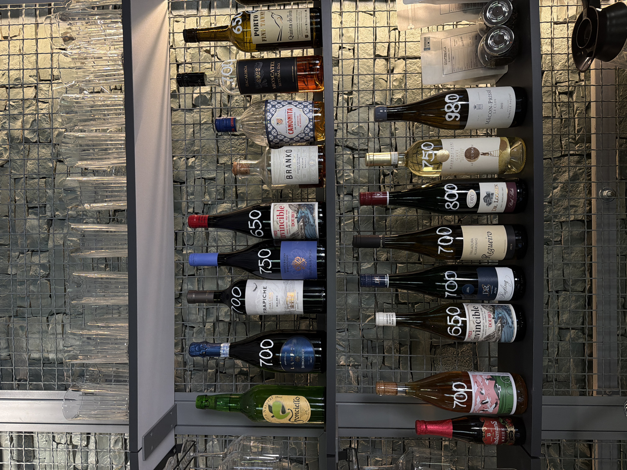 The image shows a wine rack with a variety of wine bottles. There are several rows of wine bottles, stacked horizontally on the rack. Each bottle has a label indicating the brand, type of wine, and year. The wine rack is mounted against a wall made of stone blocks. On the left side of the wine rack, there are empty glasses. There are also some coffee-related objects at the top of the right side. Based on the wine labels, the scene is likely located in a wine shop or restaurant that serves a selection of European and possibly South American wines. The location in the city of Belgrade, Serbia, is not definitively determinable from the image alone, but given the array of wines, it's probable that it is a specialized store or an upscale eatery in the area.