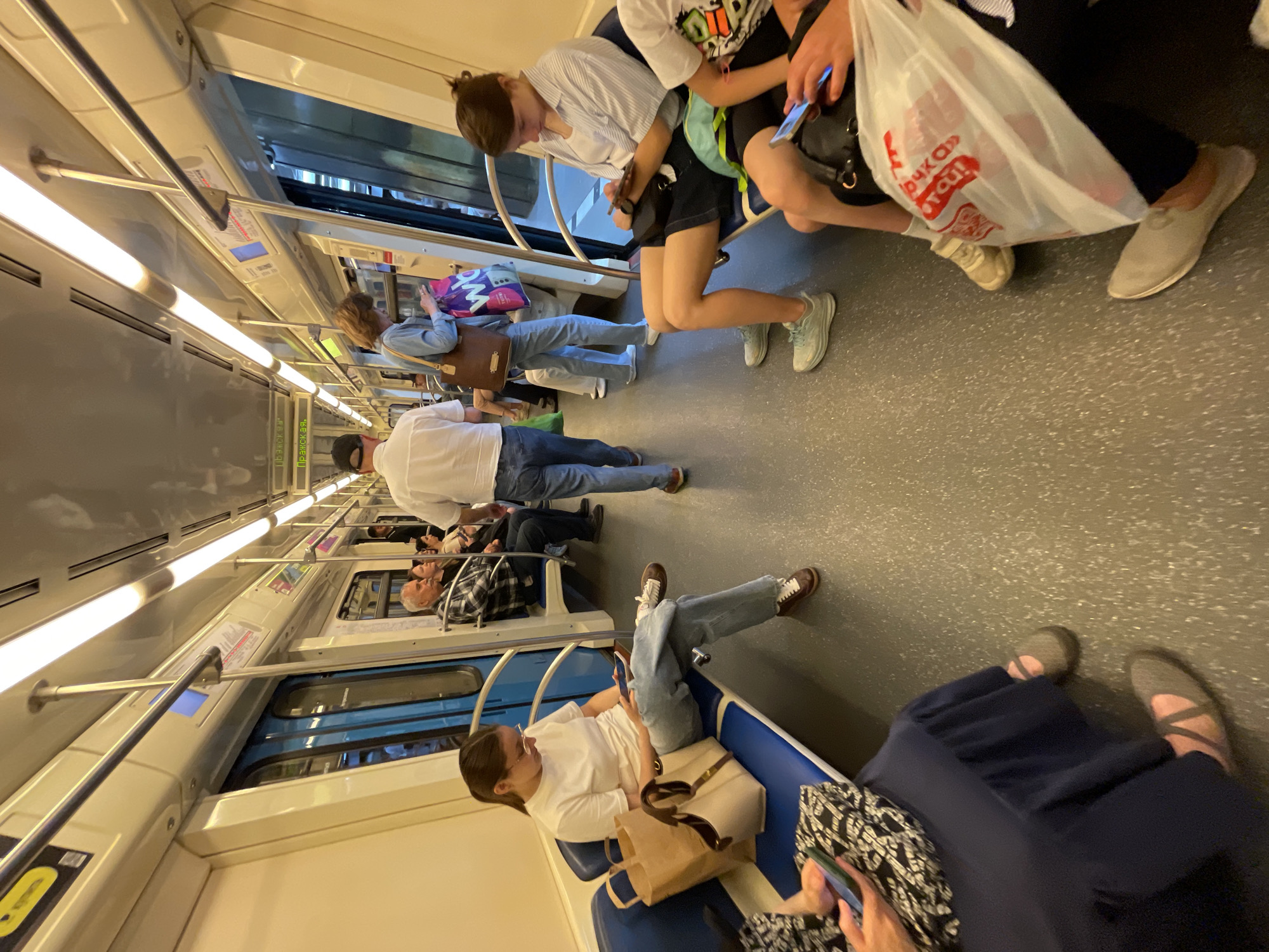 The image depicts the interior of a modern Moscow Metro train car, specifically on the Serpukhovsko-Timiryazevskaya Line, indicated by the electronic display showing "Пражская" (Prazhskaya) station. Bright, linear fluorescent lights illuminate the cream-colored ceiling and wall panels. Blue bench seating lines both sides of the car, with passengers either seated or standing in the central gray-speckled aisle.
Approximately ten individuals are visible. On the left, a young woman in a white t-shirt and jeans sits with her legs crossed, looking at a mobile phone. Beside her is a beige tote bag and a brown paper bag. In the foreground on the left, a person in a dark blue skirt and gray ballet flats is also engrossed in a mobile phone.
On the right side of the aisle, an older man in a plaid shirt is seated. Further down the aisle, a woman in a denim shirt stands, looking down, holding a brown shoulder bag and a purple "WB" (Wildberries) shopping bag. A man in a white short-sleeved shirt and blue jeans walks away from the camera down the aisle. Closer to the foreground on the right, a young woman in a striped shirt and dark shorts sits, looking at her phone, wearing light blue sneakers. Another individual in the immediate right foreground holds a white plastic shopping bag with red Cyrillic text ("дочка сын" - daughter/son) and a mobile phone. The blue train doors, some reflecting the interior and others showing glimpses of a passing platform or tunnel, are visible along the sides. Vertical and horizontal metallic poles provide handholds for standing passengers.