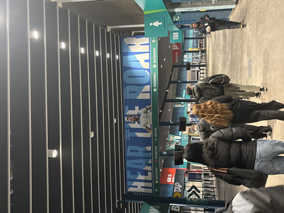 The image captures an indoor concourse or hallway within a large venue, identified as Wembley in the United Kingdom. The ceiling features dark, parallel panels with integrated lights, casting an artificial glow consistent with an evening or night setting.
Along one wall, a prominent blue banner displays the phrase "HEAR THE ROAR" in large white letters, alongside an image of a male soccer player in a white jersey. Below this, various green directional signs indicate services such as "PROGRAMMES," "MERCHANDISE," "↑ WATER POINT," "↑ INFORMATION," and "+ FIRST AID." A red sign reads "HERE TO TAP," likely for a payment or water station, while another green sign shows a female restroom symbol and the number "108," possibly indicating a gate or section.
Several people are visible, primarily from behind, suggesting movement through the concourse. In the foreground, individuals wearing dark jackets and casual clothing are walking. Further back, more people are seen near what appear to be kiosks or service counters. One person with long, wavy, reddish-brown hair is prominent among the crowd. In the distance, near the center-right, a person in a dark jacket stands, possibly a staff member, observing the flow of people.
The scene depicts attendees at a large public event, most likely a sporting event given the context of Wembley and the "HEAR THE ROAR" banner with a soccer player. The various service signs confirm the operational nature of the venue during an event.