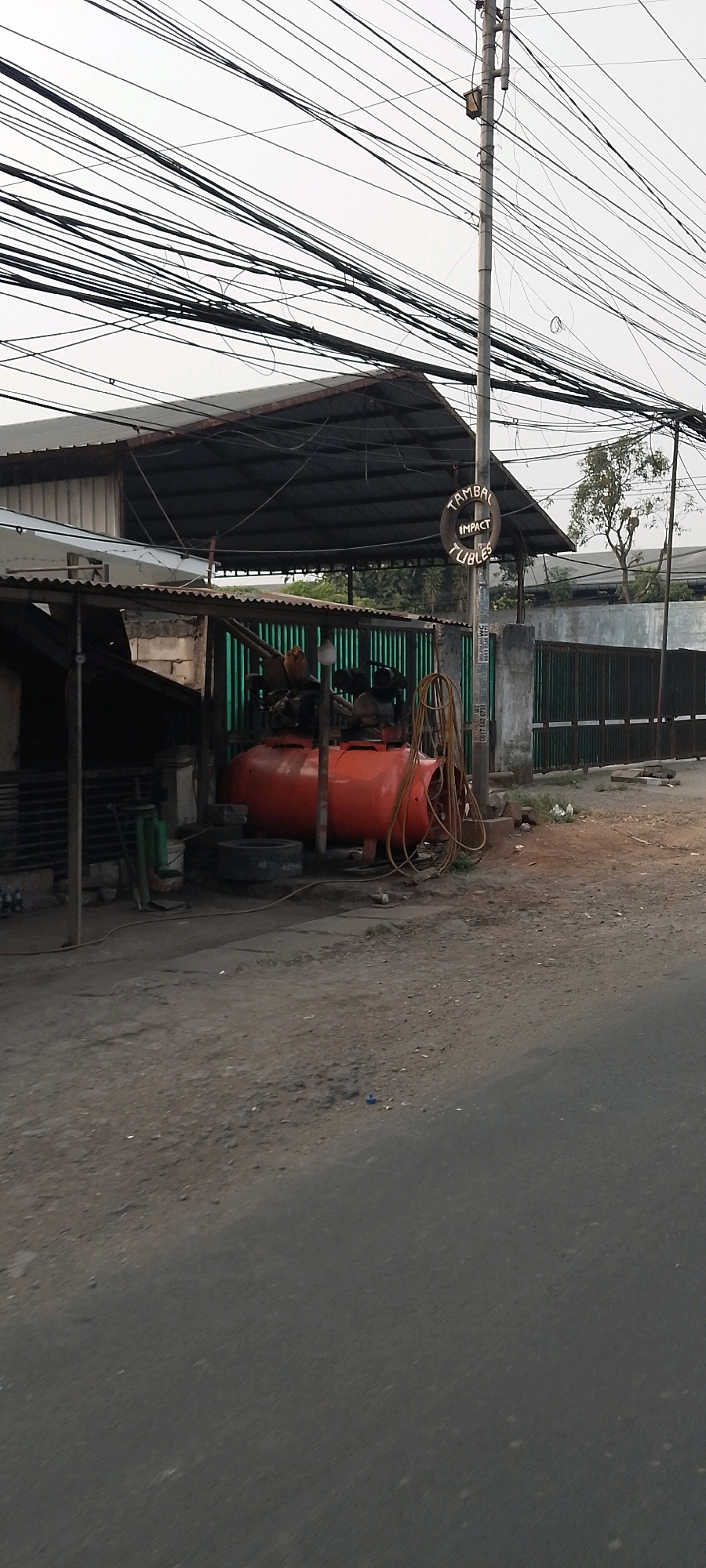 The image shows a tire repair shop in Bekasi, Indonesia. A large red air compressor is visible, along with several tires and hoses. A sign on a pole reads "TAMBAL IMPACT TUBLES," indicating tire repair services. The shop appears to be outdoors, with a corrugated metal roof providing some shelter. Overhead, a dense network of power lines crisscrosses the sky. The ground is a mix of dirt and asphalt. In the background, a green metal fence and some trees can be seen. There are no people clearly visible in the image.