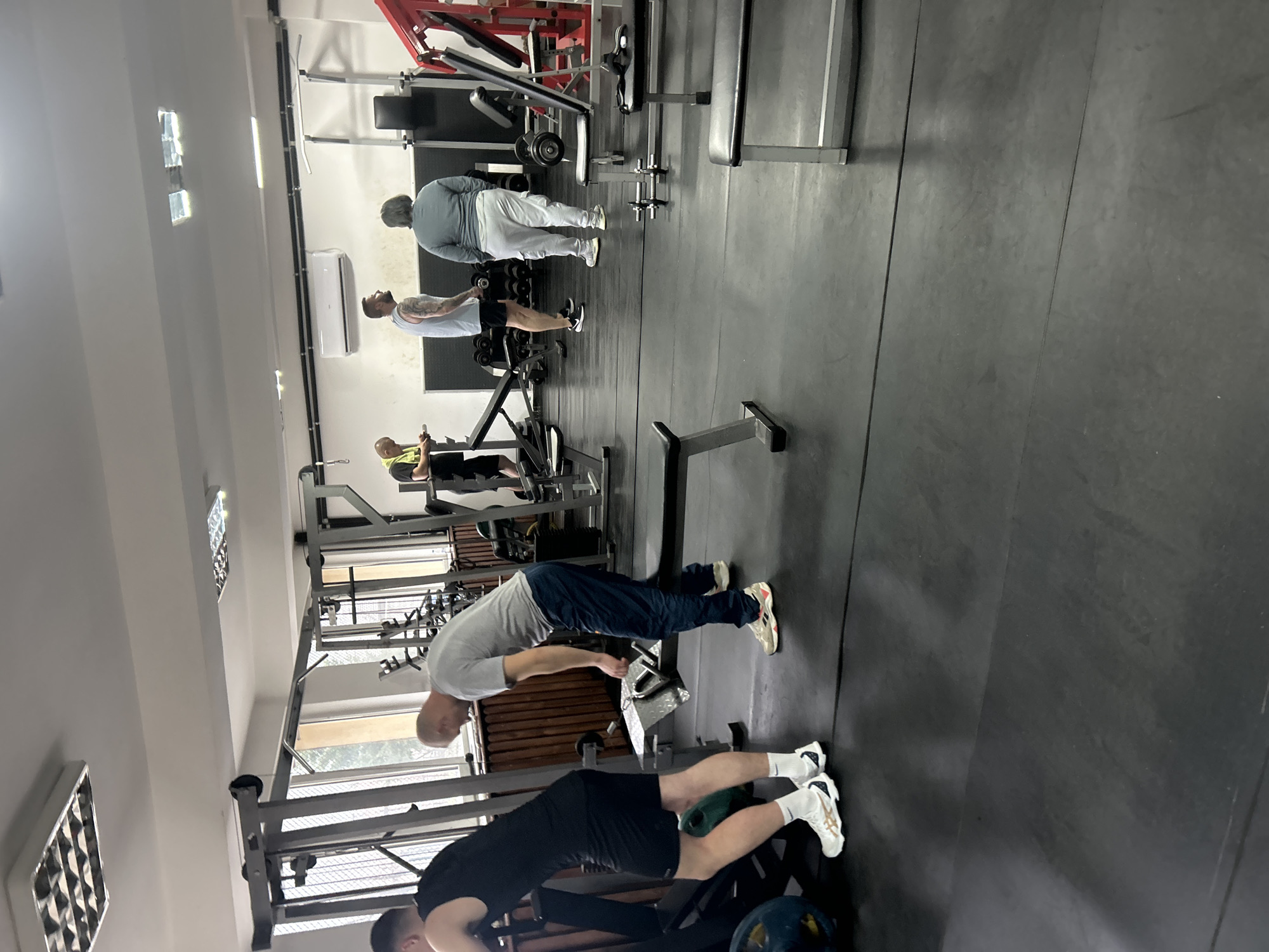 The image depicts the interior of a gym. Several individuals are engaged in weightlifting and exercise activities. The floor is made of black rubber. Weightlifting equipment is visible throughout the space. The location is presumably in Zlatograd, Bulgaria.
