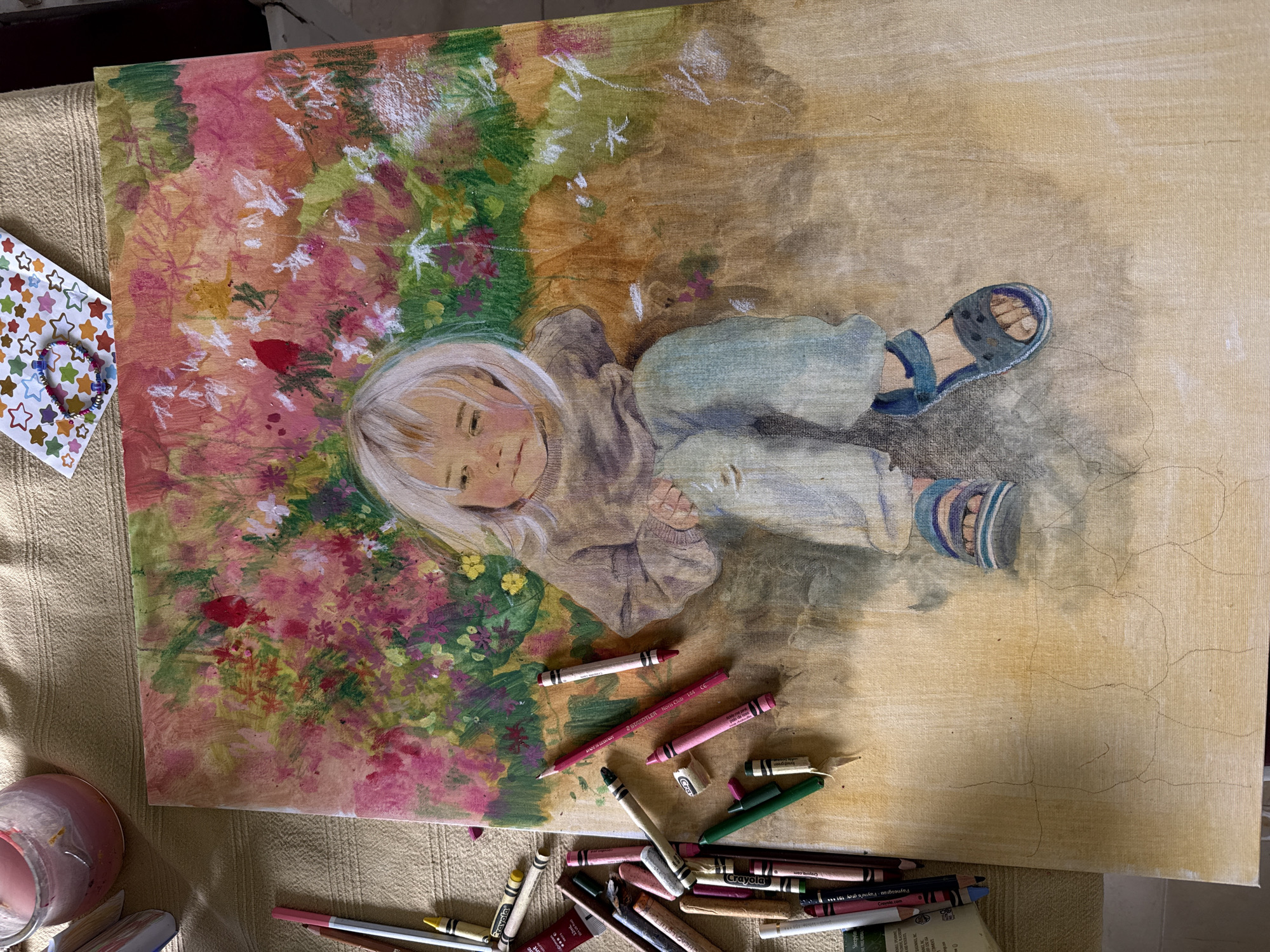 The image depicts a partially completed painting of a young child lying amongst flowers. The child has fair hair and is wearing a tan-colored shirt, blue pants, and blue sandals. The background behind the child's head is a colorful mix of pink, red, green, and white flowers. The area around the child is painted tan. The canvas is cracked with several brown lines and there are crayons scattered on the lower portion of the canvas and to the sides. In the upper left corner, there is a sheet of stickers with multi-colored stars and a bracelet made of colored beads. There is a jar of pink liquid in the lower left corner. The location of the image is unknown, it is unlikely that the image was taken in Kfar Vradim, Israel.