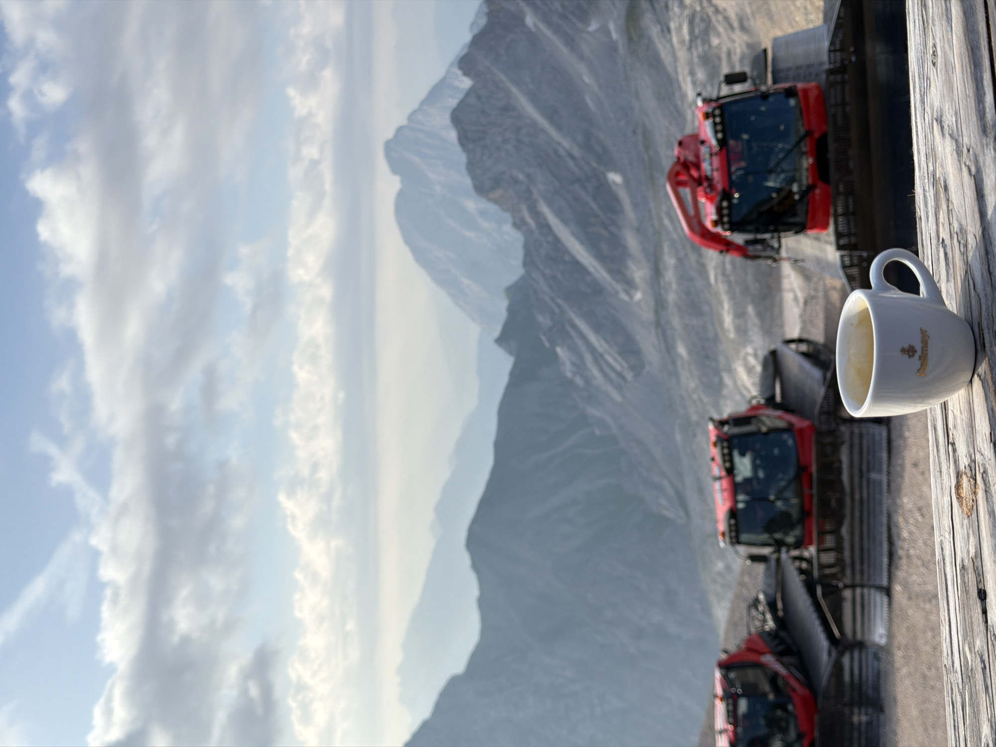 A white coffee cup, with a gold logo that reads "Dalmayr", sits on a wooden surface in the foreground. Behind the cup, a row of three red snowcats are parked. The furthest snowcat is partially obscured by the middle one, and the closest snowcat is mostly obscured by the middle one. The snowcats are all facing to the right. In the background, a vast mountain range stretches out under a cloudy sky. The mountains are layered in shades of blue and gray, with some snow visible on the peaks. The scene appears to be located at a high altitude, possibly in the Bavarian Alps, near Garmisch-Partenkirchen, Germany.