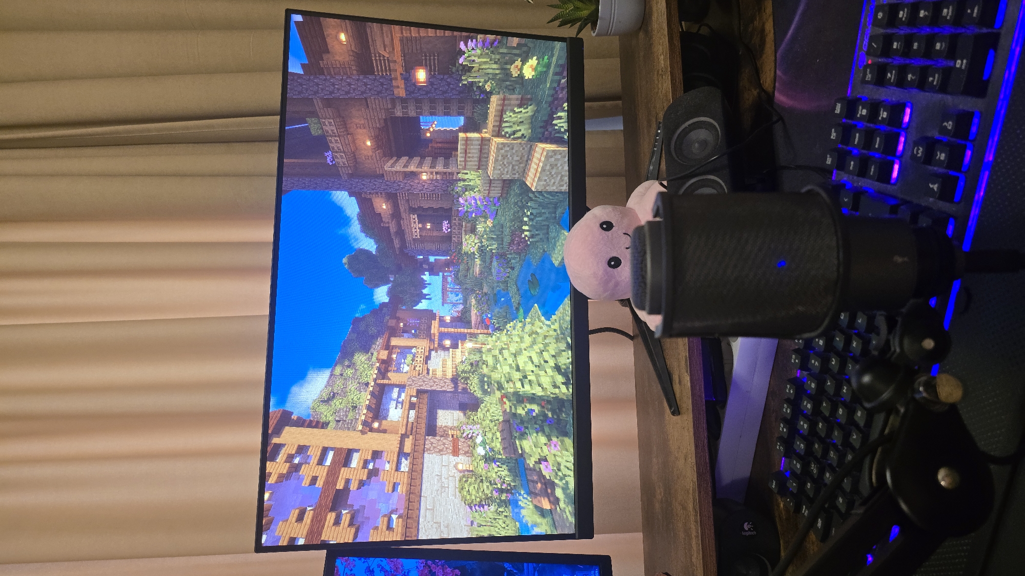 This image captures a desktop computer setup, likely configured for gaming or content creation, indoors. The central focus is a large, widescreen monitor displaying a detailed, pixelated scene from a video game, highly suggestive of Minecraft. The game environment features elaborate wooden structures, lush green foliage, colorful flowers, and a body of water under a clear blue sky, presenting a rich, custom-built world.
To the right of the monitor, a black condenser microphone with a pop filter is prominently positioned on a stand, showing a subtle blue indicator light. A small, pink plush toy with black eyes peeks out from behind the microphone and in front of the monitor, adding a personal touch. Further to the right, a gaming keyboard with striking blue and pink backlighting is visible, alongside a partially visible mouse, which bears the "Logitech" brand name. Two compact black speakers are also part of the setup, one situated beside the monitor and another near the keyboard.
In the upper right portion of the frame, a small potted succulent plant is visible. The entire setup rests on a wooden desk surface. On the far left, light-toned curtains, possibly beige or cream, are drawn. The arrangement of the microphone, gaming keyboard, and monitor suggests an environment designed for interactive media consumption or creation, such as gaming, streaming, or voice recording. The time of day is not explicitly indicated, and there are no direct visual cues within the image to specify its location in Sofia, Bulgaria.