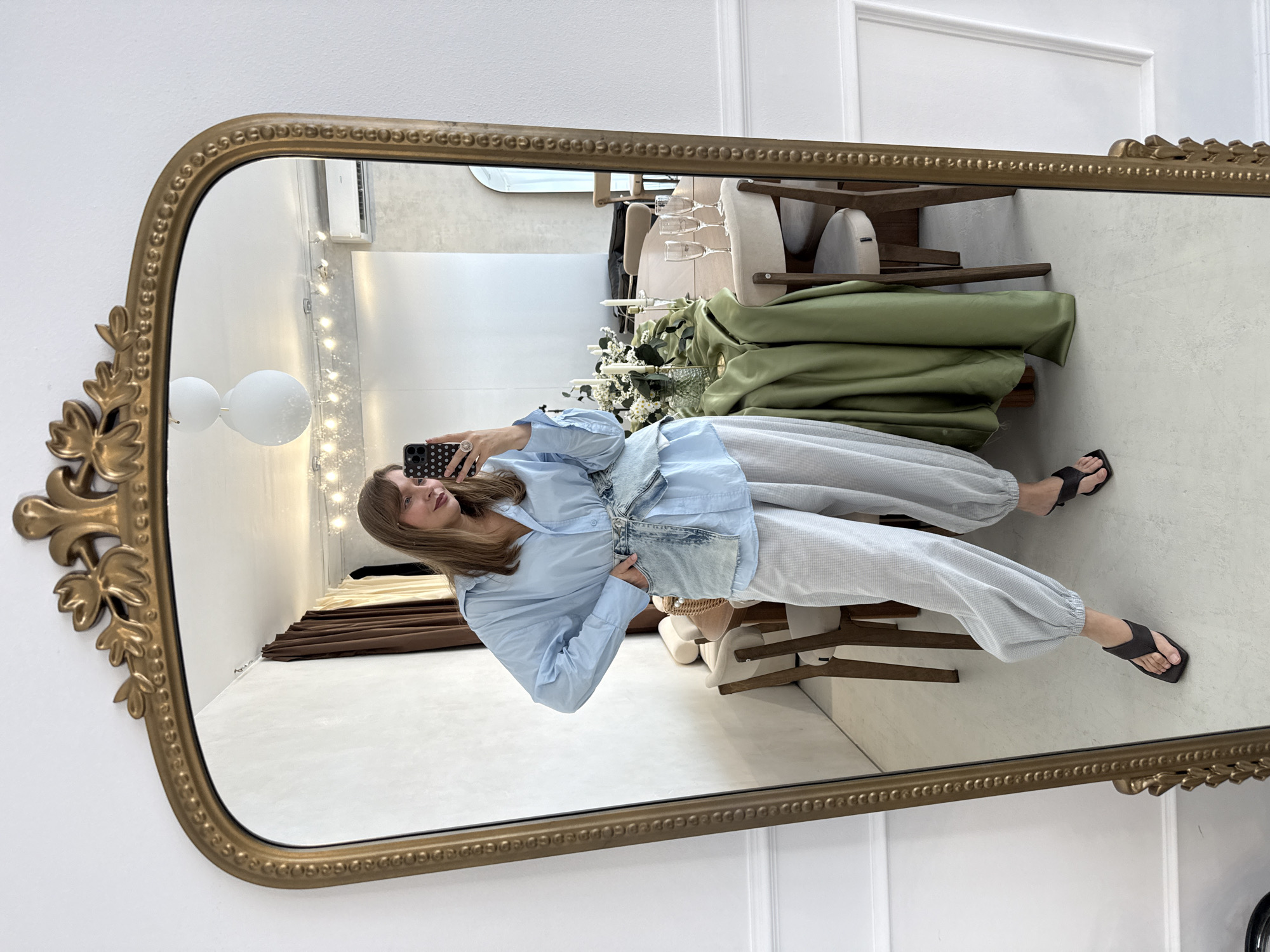 The image is a full-length mirror selfie taken indoors. The subject is a person in casual attire, wearing a light blue oversized shirt, a denim vest, and light grey sweatpants, paired with black flip-flops. They are holding a black smartphone with a white speckled phone case, taking the picture. The mirror has an ornate, gold-colored frame with decorative leaf motifs.
The reflection in the mirror shows a room with white walls and some decorative elements. In the background, there are stacked green fabric drapes, a table with glassware and candles, and part of a wooden chair. A string of fairy lights is visible on the wall behind the subject. The lighting suggests it could be daytime, but the indoor setting makes it difficult to determine the exact time of day or weather. The overall ambiance is casual and relaxed. No visible text is present in the image. The location context provided is Izhevsk, Russia.