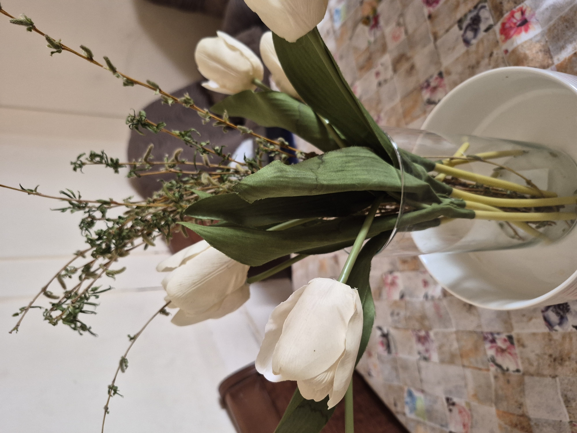 A still-life arrangement of artificial flowers and greenery is depicted in an indoor setting. The main subjects are several cream-colored artificial tulips with prominent green leaves and stems, accompanied by sprigs of artificial greenery featuring thin brown stems and small, light green, bud-like foliage, possibly mimicking thyme or a similar plant. These are arranged in a clear glass vase, which is partially visible and appears to be placed within a larger, white, cylindrical container or pot.
The vase and flowers rest on a patterned surface, likely a tablecloth or placemat, composed of square or rectangular patches in various muted colors including beige, brown, pink, and light blue. In the blurred background, there are indications of a light-colored wall or surface, as well as dark upholstered furniture and a piece of dark wooden furniture.
The image is oriented on its side, with the vase and flowers appearing horizontally. There are no people present, no specific activity or event taking place, and no visible text. The lighting suggests an indoor setting, but the time of day and weather are not discernible. The location context provided is Yerevan, Armenia, though no visual cues within the image confirm this.