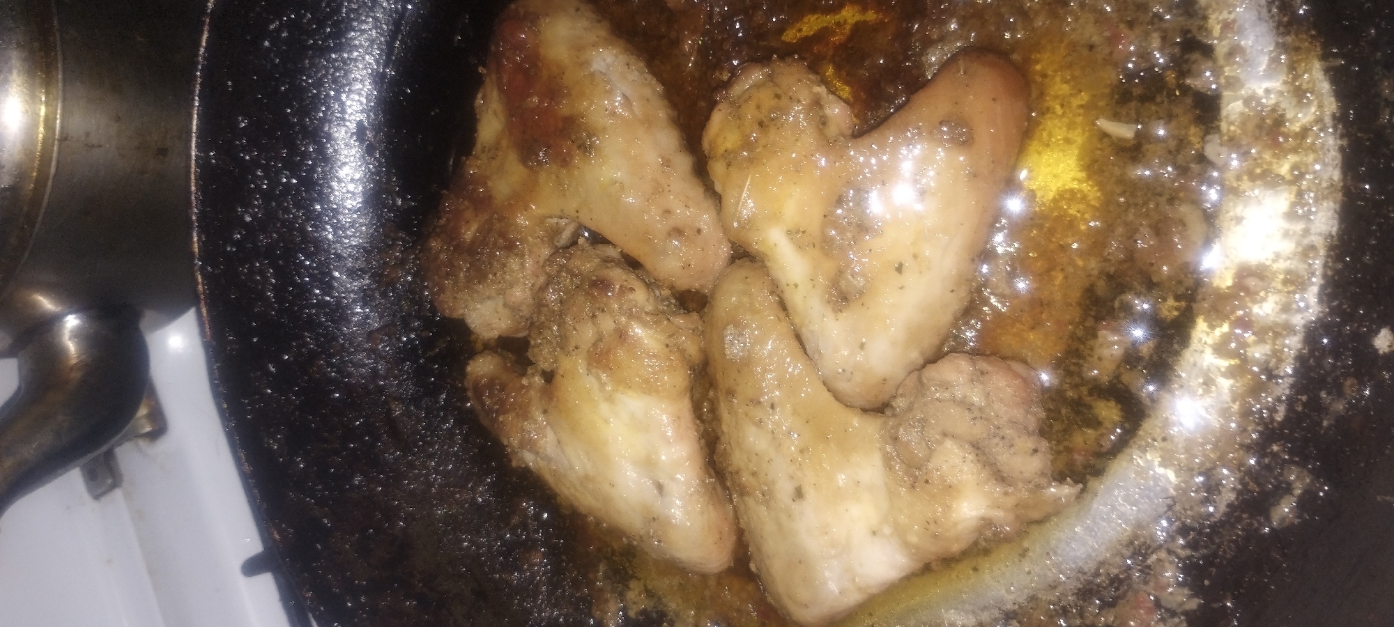 Chicken wings are being fried in a dark pan with oil. The wings are seasoned and appear to be cooked. The pan is on a stovetop, and a kettle is visible in the background. The scene is an indoor kitchen.
