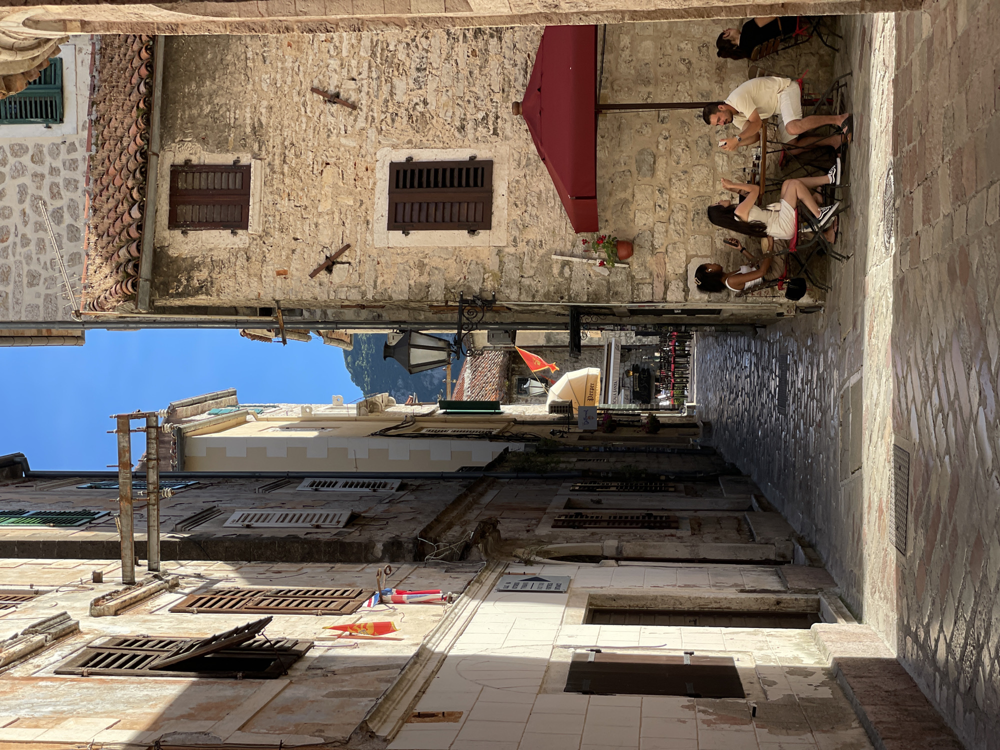 The image depicts a sun-drenched, narrow cobblestone alleyway in the historic Old Town of Kotor, Montenegro. The architecture features ancient stone buildings with weathered facades, wooden shutters, and terracotta tiled roofs. On the left, a building shows peeling paint and flags, one of which is red with a yellow emblem. On the right, a stone building has a dark wooden shuttered window and a red umbrella shading a small outdoor cafe.
Seated at a table under the umbrella are three people. A woman in a white tank top and skirt, wearing earbuds, looks at her phone. To her right, a woman in a white dress is seated, holding a fork and looking at her phone. A man in a pale yellow polo shirt and white shorts sits opposite the woman in the white dress, also looking at his phone and holding a beverage. Further to the right, another person with dark hair is partially visible sitting at a separate table.
The alleyway is paved with uneven cobblestones, and the sunlight casts strong shadows. In the distance, the alley continues, revealing more buildings and hinting at the medieval character of Kotor. The overall scene suggests a leisurely afternoon in a charming, historic European town.