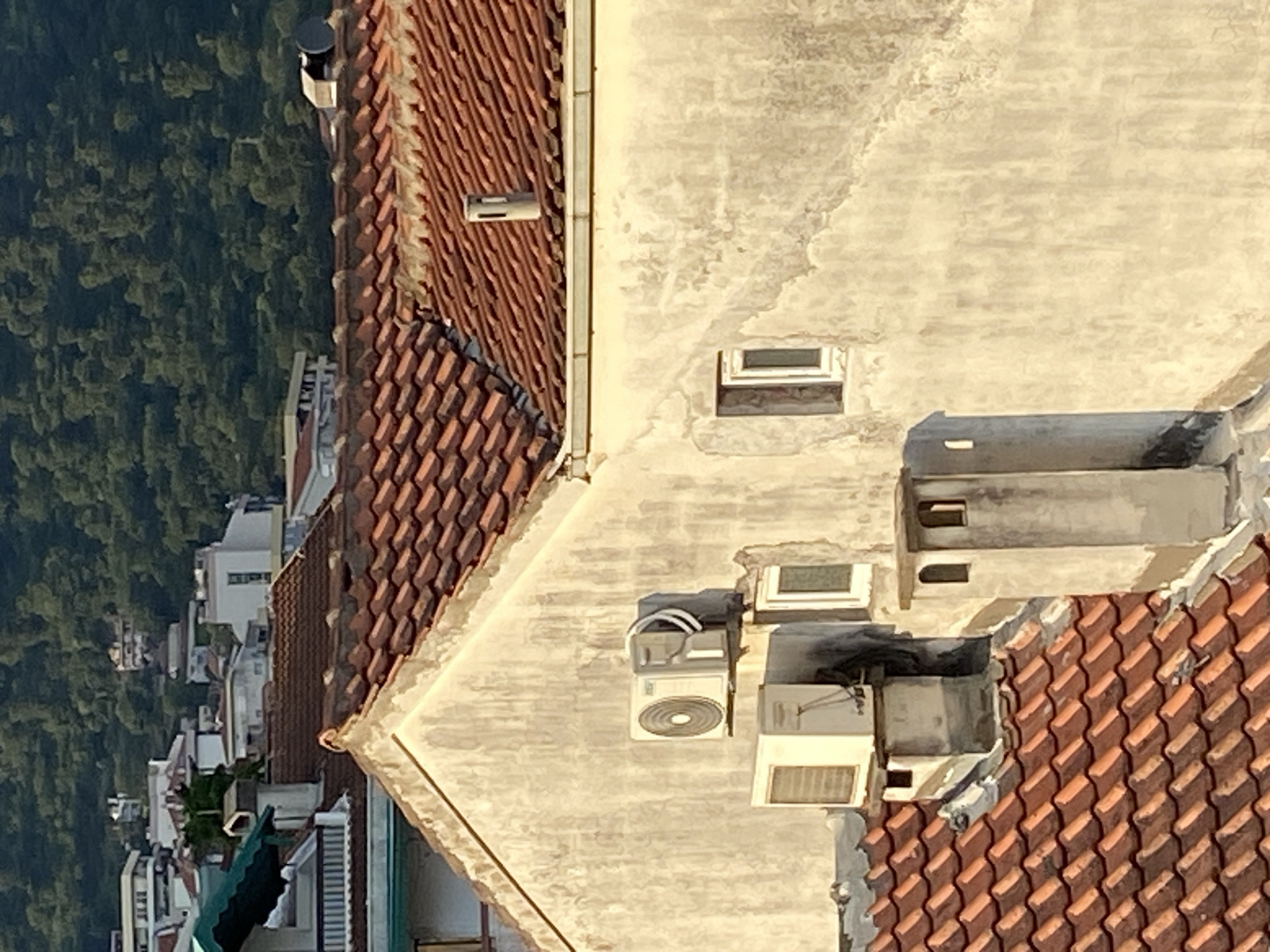 The image depicts an aerial view of rooftops in Budva, Montenegro. The scene is dominated by terracotta-tiled roofs and light-colored concrete structures. Several air conditioning units are visible on the roofs, as well as small windows. In the background, a dense green forest can be seen, juxtaposing the urban environment. The image suggests a residential area with buildings in close proximity.