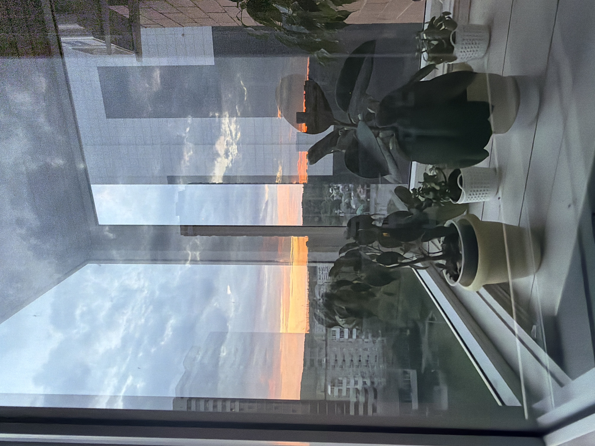 The image captures a serene view from a window during either sunrise or sunset, looking out over an urban landscape. Several potted plants are arranged on the windowsill, casting dark silhouettes against the vibrant sky. The sky is a prominent feature, displaying warm hues of orange, yellow, and pink near the horizon, gradually fading into lighter blues and grays higher up. Scattered clouds are visible, both directly in the sky and as reflections on the windowpane.
The window glass creates a layered effect, with reflections of the clouds, the indoor plants, and possibly parts of the room's interior overlapping the exterior scene. In the distance, numerous buildings of varying heights can be seen, indicating an urban or suburban setting. The overall atmosphere is calm, highlighted by the soft, warm light of the early morning or late evening. No specific activity is taking place, nor is any text visible. The location is an unknown urban area.
