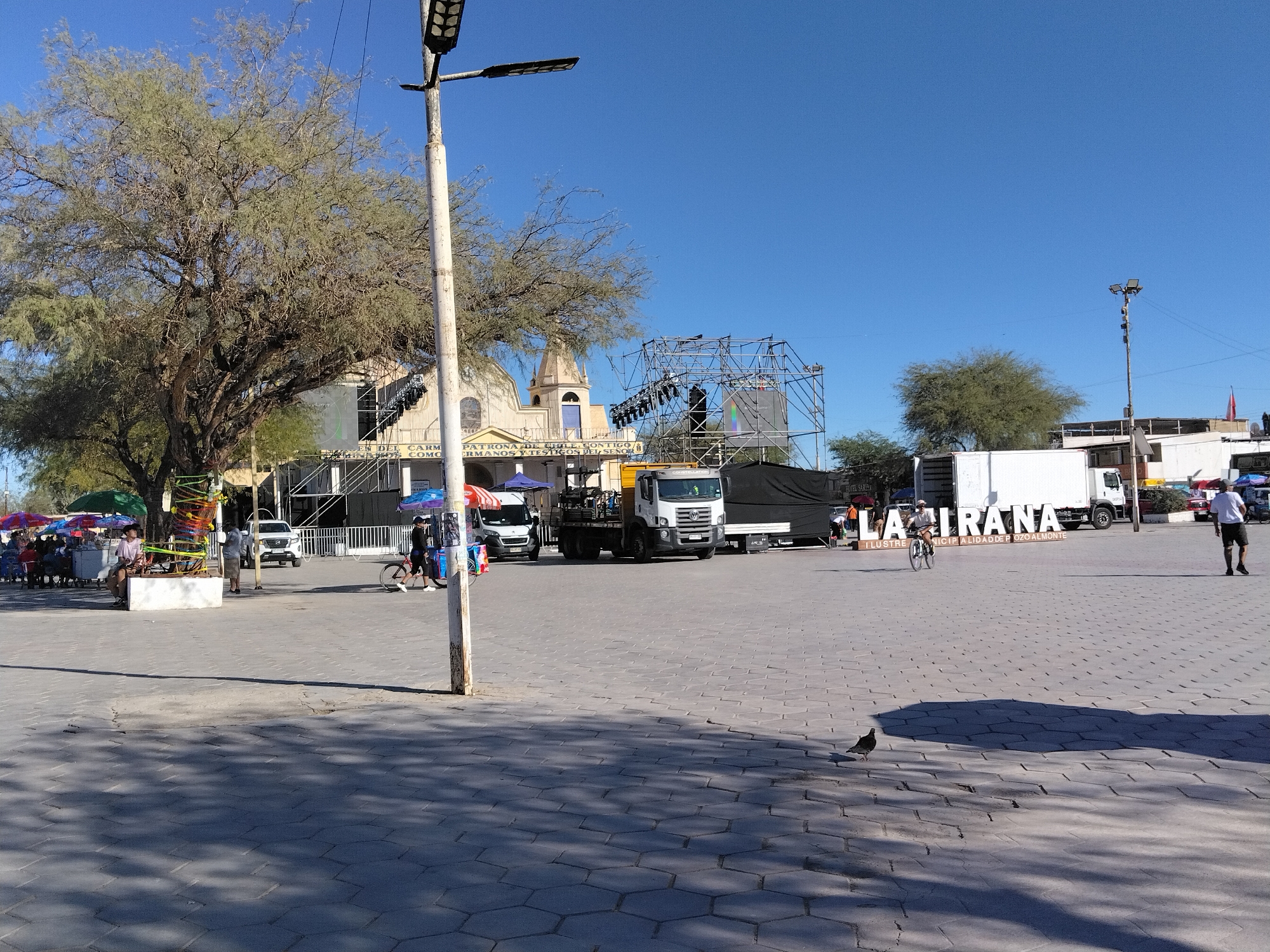 This outdoor scene captures a public square in Pozo Almonte, Chile, bathed in bright sunlight under a clear blue sky, suggesting it's daytime. The foreground is paved with hexagonal stones, with long shadows cast by unseen objects, indicating a sunny day.
The main subjects include a large, mature tree with sparse foliage on the left, and a white building resembling a church or municipal hall in the background, adorned with inscriptions like "PATRONA DE CHILE CONTIGO" and "CRMANOS Y TIGOS DEL SEÑO." A large stage structure with scaffolding and lighting is set up in front of this building.
Several vehicles are present, including a white truck, a smaller white van, and a pickup truck. People are scattered throughout the square, some browsing stalls with colorful umbrellas, others walking, and one person is riding a bicycle. A prominent sign with large white letters spelling "LAATRANA" stands in the middle ground, with smaller text below that reads "ILUSTRE MUNICIPALIDAD DE POZO ALMONTE," confirming the location. The overall atmosphere appears to be that of an outdoor event or market day. A bird is visible in the foreground on the pavement.