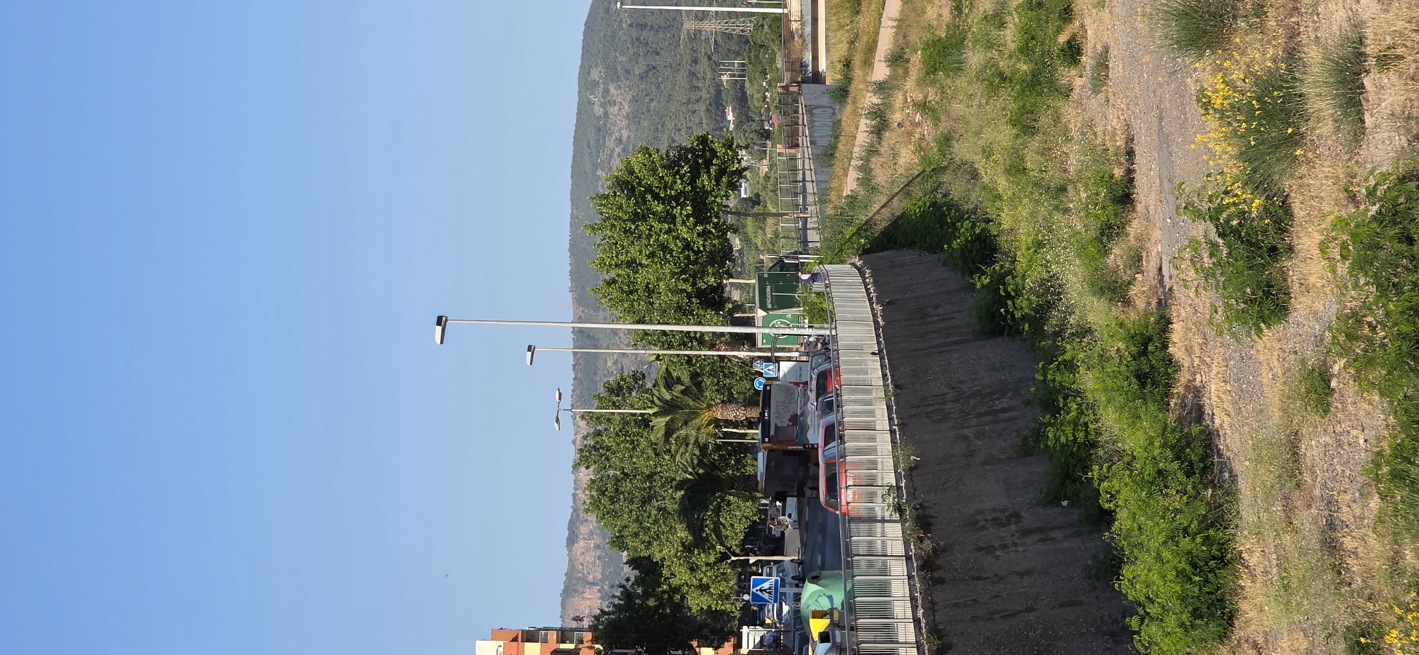 The image depicts a sunlit outdoor scene, likely a road or street running alongside a natural hillside, with mountains in the background. The sky is a clear, bright blue, indicating good weather, possibly daytime in the morning or early afternoon.
In the foreground and midground on the left side, there's a paved road or urban area. Several modern street lamps with rectangular fixtures are visible, lining what appears to be a street or promenade. Lush green trees, including at least one palm tree, are planted along this area. Partially visible at the very bottom left are parts of multi-story buildings with orange and white facades. Several vehicles, including cars (one red) and what looks like a bus or large truck, are present, suggesting an active street. A prominent green container or truck with the word "MERCADONA" clearly visible on its side is parked or positioned on the left. Blue square road signs, one appearing to indicate a pedestrian crossing and another with an arrow, are also present. A metal railing separates the urban side from a lower concrete path or retaining wall.
On the right side, a natural, gently sloping or steep hillside covered in dry grass, green bushes, and scattered yellow wildflowers is prominent. Dirt patches and possibly eroded areas are visible on this incline. A concrete path or set of steps runs along the base of this hillside, next to the metal railing.
In the background, a large, tree-covered mountain range dominates the horizon under the clear sky. Power lines and support pylons can be seen against the mountain on the upper right side, suggesting infrastructure in the distant landscape. There is one person visible near the green Mercadona container, but their activity is not clear. The overall impression is a sunny, Mediterranean urban-natural landscape.