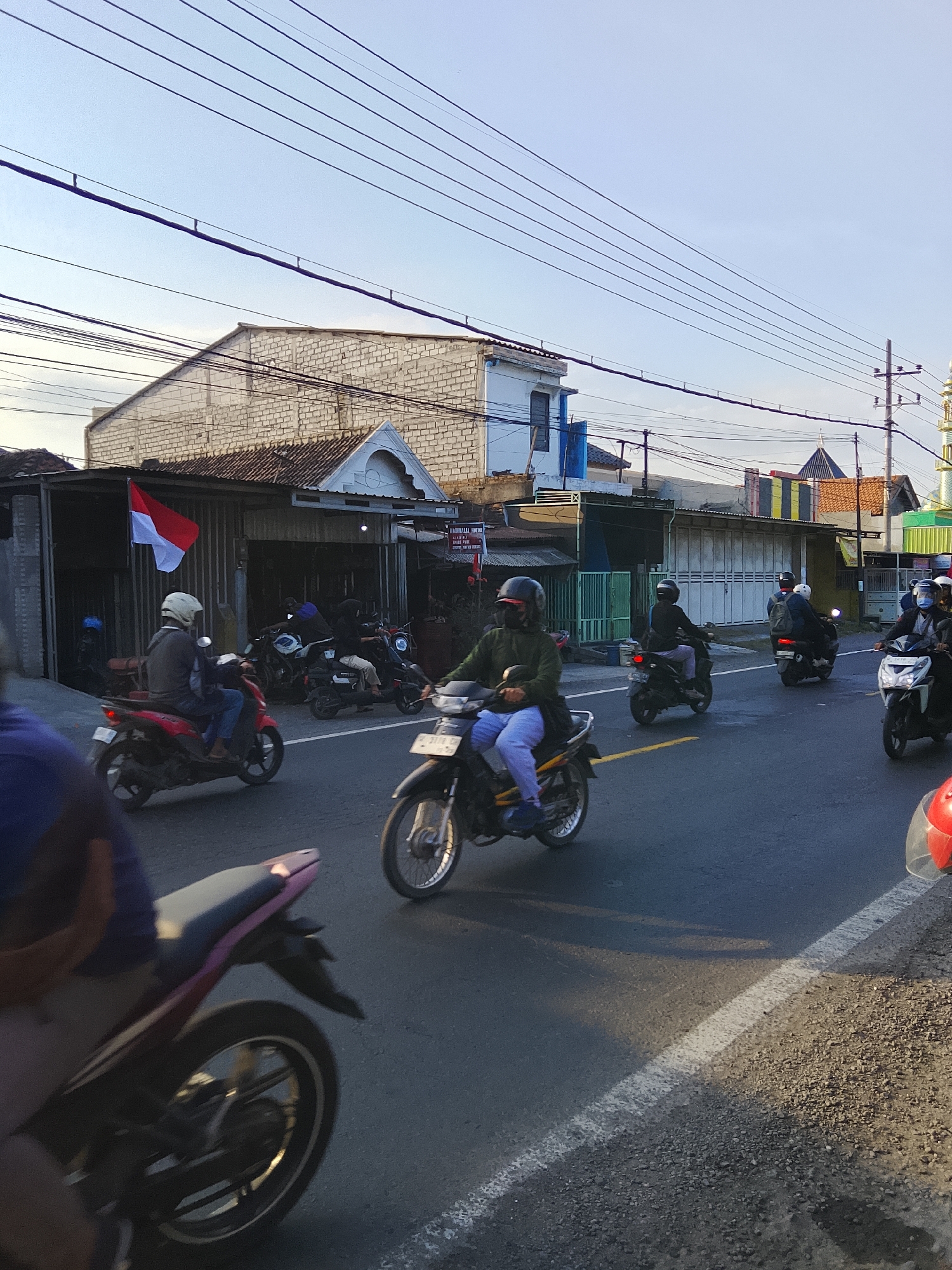 In Banjarsari, Indonesia, a street scene captures multiple motorcycles in motion. A red and white Indonesian flag is prominently displayed on the left side of the image, attached to a building. Several individuals are riding motorcycles on a paved road, some wearing helmets. The buildings lining the street appear to be commercial establishments, with one featuring a white brick facade. Overhead power lines crisscross the sky, which is a light blue. A minaret of a mosque is visible in the background on the right. The overall impression is a typical day on a busy street in a Southeast Asian urban environment.
