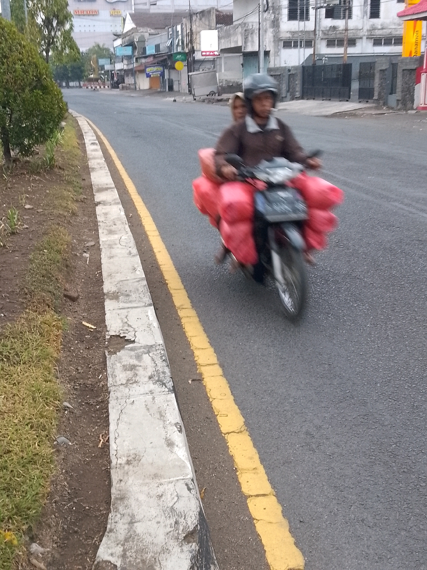 Two individuals, appearing to be an adult male and a younger person, are riding on a motorcycle. The motorcycle is laden with red, presumably fabric or soft goods, secured behind the riders. The male is driving and wearing a brown jacket and a dark helmet. The younger person is seated behind him, also appearing to wear a head covering. They are traveling on a paved road with a yellow line marking the edge of the lane. To the left of the road is a grassy embankment with a concrete curb. In the background, buildings and trees suggest an urban or semi-urban environment. The scene is captured in motion, indicated by the blur of the motorcycle and its occupants. The location is identified as Jember, Indonesia.