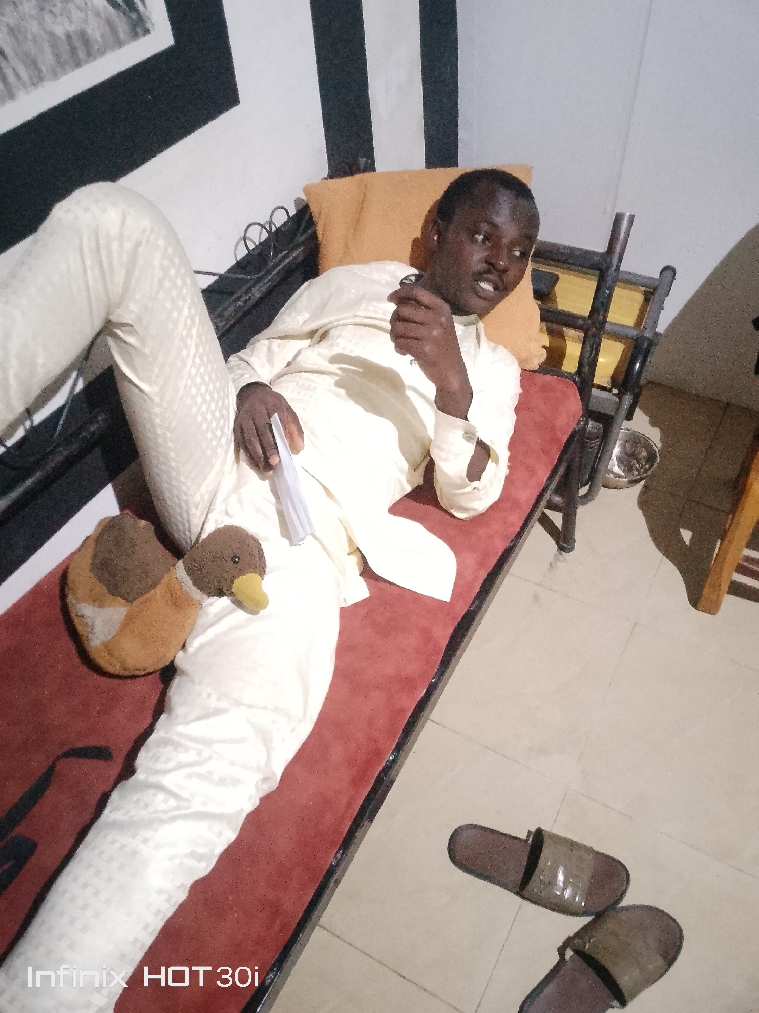 In an indoor setting, a Black male reclines on a narrow, red-upholstered daybed. He is wearing a cream-colored traditional outfit and appears to be speaking. His right hand is raised, with his index finger extended and his thumb touching his chin. A folded white object, possibly a fan, rests on his left leg. Beside his left leg, a brown and orange stuffed duck toy is placed. The daybed is supported by a black metal frame. An orange pillow is behind the man's head. In the background, a wall is painted with alternating black and white vertical stripes. On the floor to the right of the daybed, a pair of brown slippers are visible. A metallic bowl is placed on the floor to the right of the daybed. The flooring appears to be tiled. A watermark at the bottom left of the image reads "Infinix HOT 30i".