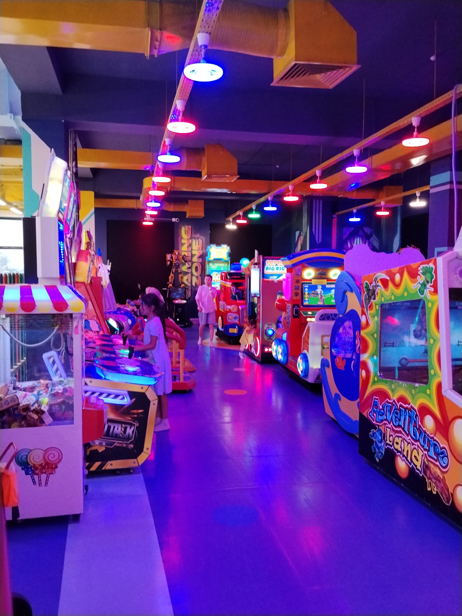 The image depicts the interior of an arcade, likely located in Bishkek City, Kyrgyzstan. The environment is dimly lit and characterized by vibrant neon lights and colorful arcade machines. The floor is a deep purple, reflecting the surrounding lights. Overhead, yellow ductwork and a dark ceiling are visible, with numerous disc-shaped lights in various colors including blue, red, and white.
In the foreground, to the left, is a large, colorful arcade machine with a striped canopy, resembling a claw machine, filled with plush toys. To its right is another arcade machine with a black and yellow design, displaying the word "TASK".
Further into the arcade, a young girl in a light-colored dress is seen playing a video game. She is facing away from the camera, engrossed in the game. Behind her, several other arcade machines are lined up. One machine is designed to look like a red train with flashing lights, and another, further down the row, is a larger, more elaborate game with bright graphics and a cartoonish design, featuring the text "Adventure Land" in colorful lettering.
In the background, a man in a light-colored shirt and shorts stands near a bright red and blue arcade game, possibly a racing simulator. Above him, a large sign reads "GAMING ZONE" in illuminated letters, with a robotic figure visible behind it. The overall atmosphere suggests a lively entertainment venue.