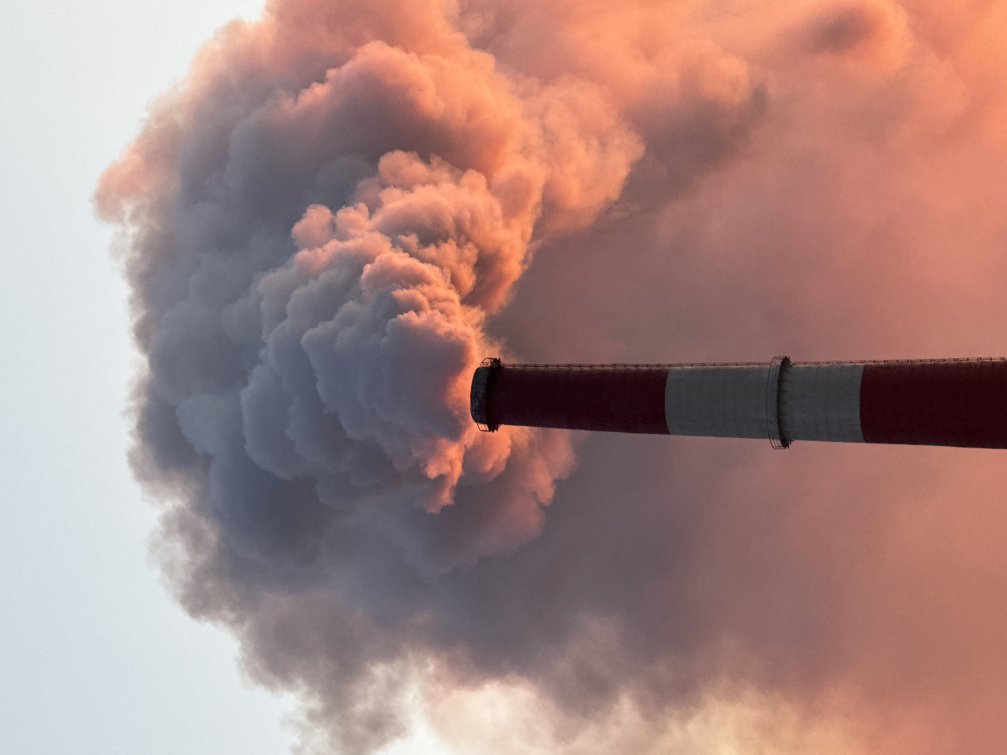 This image captures a close-up view of a tall, red and white striped industrial smokestack in Norilsk, Russia, releasing a voluminous plume of dark smoke. The smoke billows upwards and to the left, catching the warm hues of a sunset or sunrise, which casts a vibrant orange and pink glow on its edges. The sky above is a pale, hazy blue. The base of the smokestack is not visible, and the image focuses on the upper portion and the dramatic emission. There are no people or other subjects discernible in the frame. The environment is characterized by the industrial pollution spewing from the smokestack against a backdrop of a colorful sky.