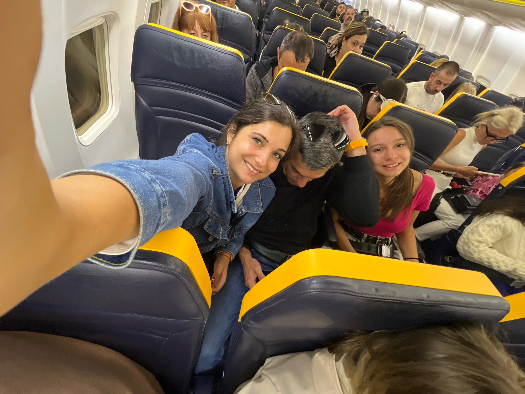 The image shows the interior of an airplane cabin, with rows of blue seats with yellow trim. Several passengers are visible. In the foreground, a young woman in a denim jacket is taking a selfie, smiling at the camera. Next to her is a man wearing sunglasses. Behind them, other passengers are seated, some looking forward, others looking down or to the side. The scene is brightly lit, suggesting interior cabin lighting. The overall impression is of a group of people on a flight. The location is identified as Uttlesford, United Kingdom, which is the location of Stansted Airport, a major hub for budget airlines that often fly aircraft of this type.