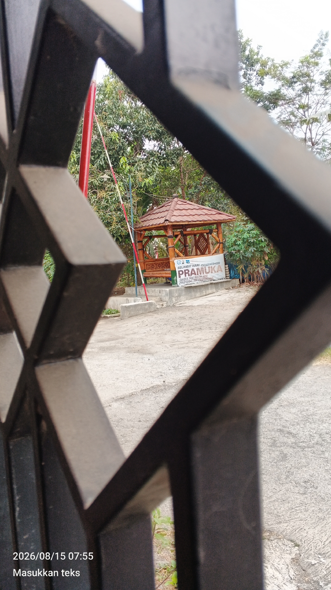 The image shows a gazebo with a brown tiled roof situated outdoors. A banner displaying "SELAMAT HARI" and "PRAMUKA" in large green lettering is visible next to the gazebo. A red and white pole is also present in the background. The scene is framed by a dark, diamond-patterned metal fence in the foreground. The ground appears to be paved. The timestamp in the bottom left corner reads "2026/08/15 07:55". The text "Masukkan teks" is also present below the timestamp. Based on the banner, the location is likely related to a Pramuka (Indonesian Scout Movement) event or facility in Jeru, Indonesia.
