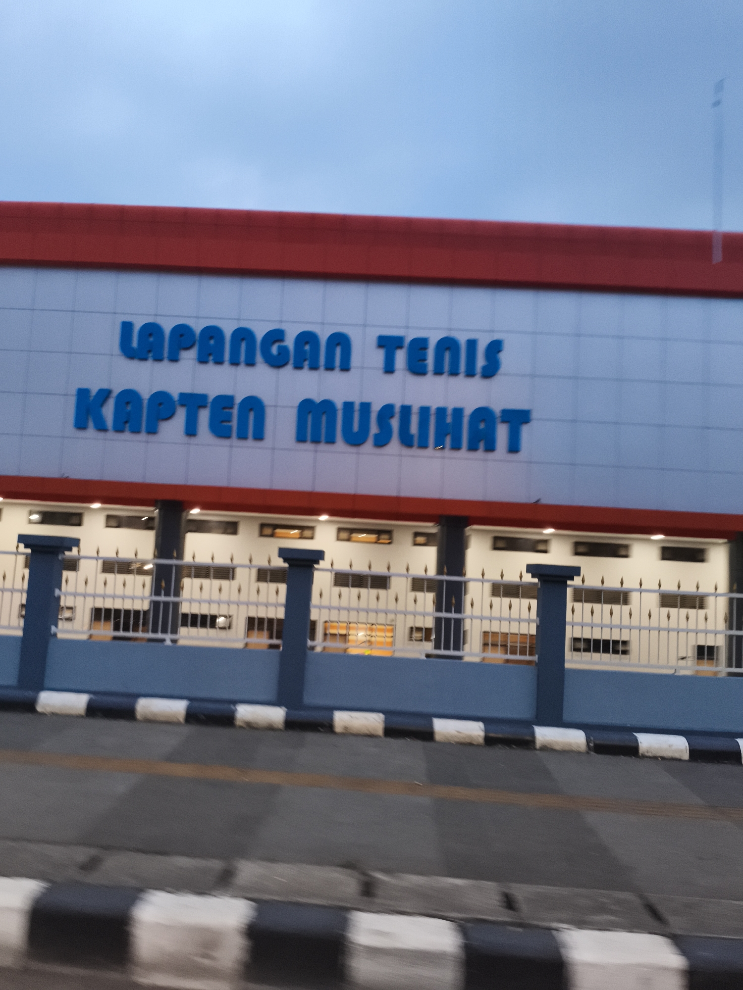 The image captures the exterior of a building, likely a sports facility, under an overcast sky or during twilight. The building features a distinctive red upper border atop light blue and white paneled walls. Large, blue, three-dimensional lettering on the facade clearly spells out "LAPANGAN TENIS KAPTEN MUSLIHAT," which translates to "Tennis Court Captain Muslihat."
In front of the building, a fence with dark blue posts and white and gold-colored vertical railings is visible. Beyond the fence, the ground floor of the building appears lit from within, with warm light emanating from windows. The foreground shows a paved surface, possibly a road, marked with a yellow line and black and white striped curbs or road barriers. The slight motion blur, especially noticeable in the foreground, suggests the photograph was taken from a moving vehicle. The overall setting points to a public or communal tennis facility located in Cibinong, Indonesia.