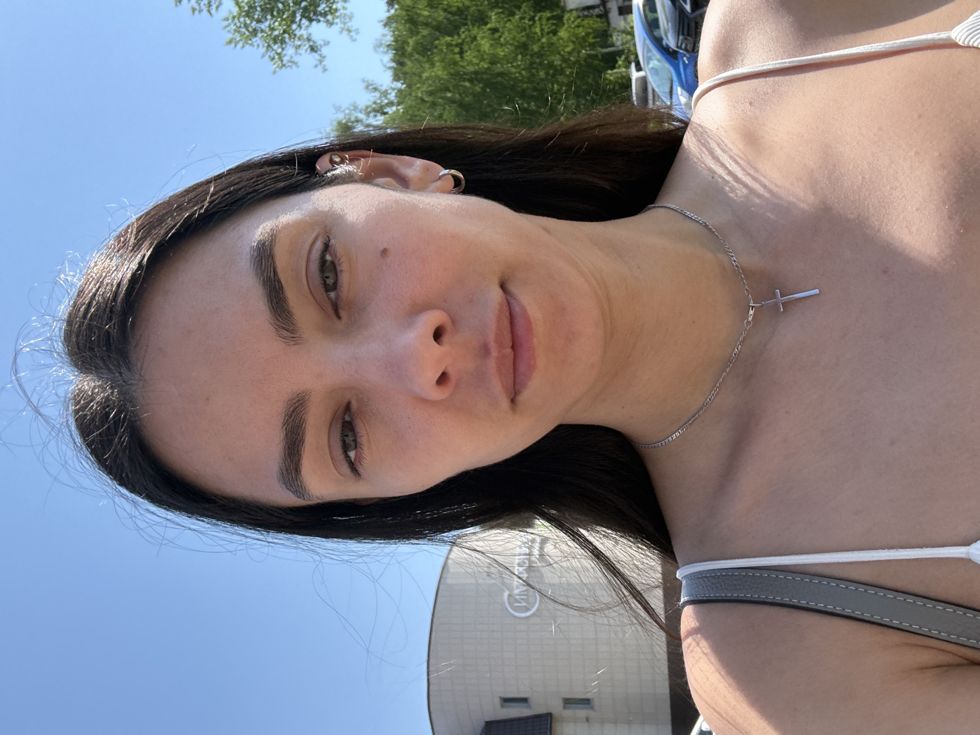 A young woman takes a selfie outdoors on a sunny day. She has dark hair, fair skin, and is wearing a white top with thin straps and a silver necklace with a cross pendant. She has multiple piercings in her left ear.
In the background, a building with a circular logo is visible. Based on the logo, the location appears to be Chaikovsky, Russia, likely near or in the vicinity of the "Tchaikovsky" sports complex. Other elements in the environment include a blue sky, trees, and a vehicle.