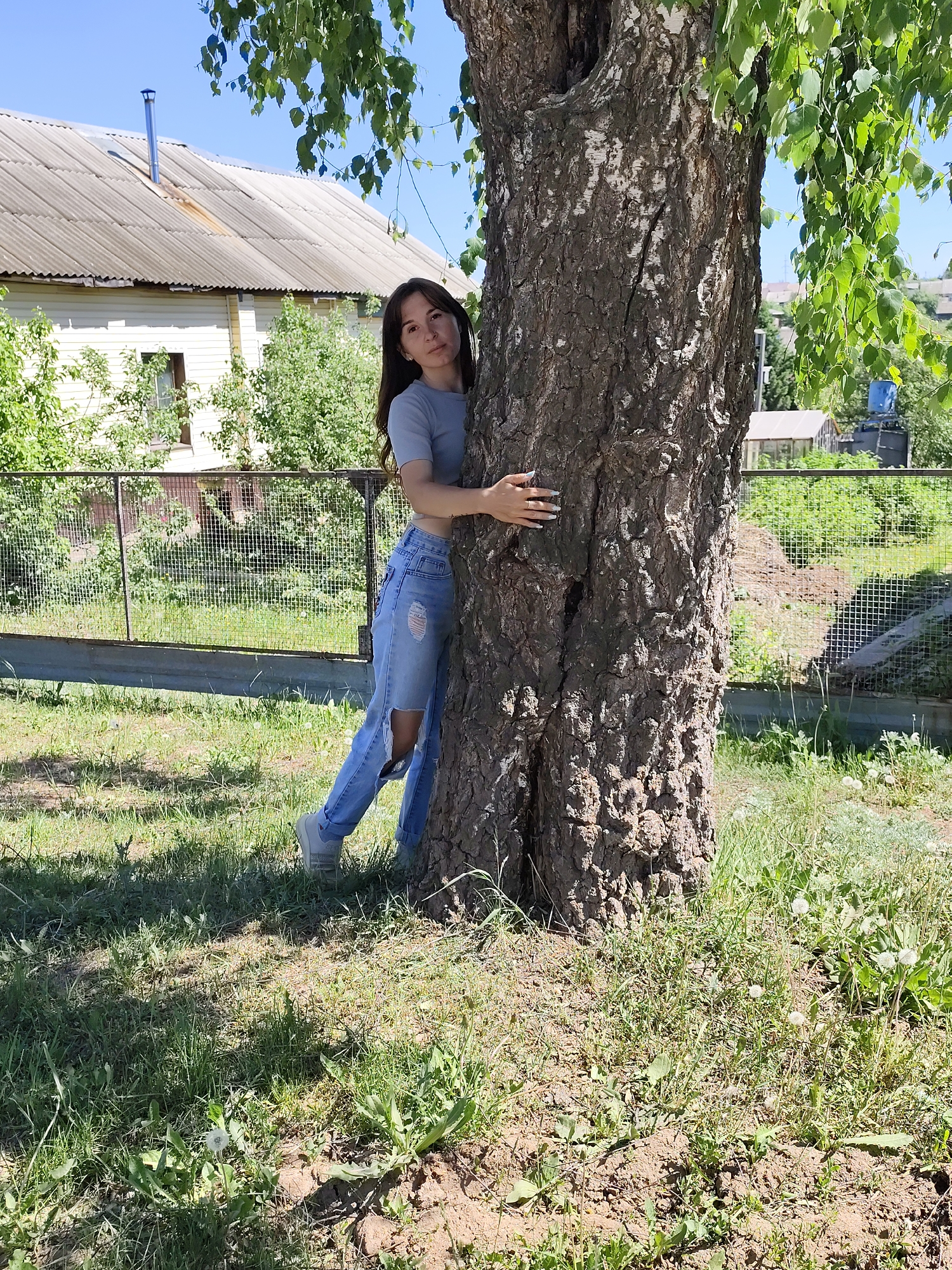 A woman is standing next to a tree in Фоки, Russia. She is wearing a light gray cropped top, ripped blue jeans, and gray sneakers. She has long brown hair and is looking at the camera. She is leaning against a tree, with a white house, a metal fence, green trees, and a blue sky in the background. The tree has thick, textured bark and green leaves. The ground is covered in grass and some dandelions.