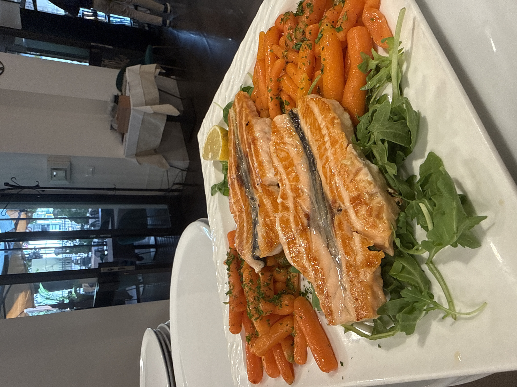 This is a close-up, eye-level, indoor, natural light, medium-shot photograph of a dish of grilled salmon and glazed carrots. The salmon is served in three portions, each cut into thick fillets, with a crispy, golden-brown exterior and a flaky pink interior. The salmon is placed on a bed of fresh, green arugula. The carrots are small, baby carrots, glazed and seasoned with chopped parsley. A lemon wedge is positioned on the arugula next to the salmon. The dish is served on a large, white, rectangular platter with a textured rim.
In the background, out of focus, is a restaurant setting. The environment appears to be a dining area with tables covered in white tablecloths and chairs. Through a glass doorway, an outdoor seating area with greenery and awnings is visible. The lighting is soft and diffused, creating a warm and inviting atmosphere. The overall impression is one of a delicious and elegantly presented meal.
The image's subject matter and presentation suggest it was taken in a restaurant or a similar dining establishment. The presence of multiple tables, tablecloths, and patrons in the background points to a public eatery. Without further information or context, it is not possible to definitively identify the city of Trento, Italy, or any specific location within it. However, the dish itself is typical of upscale Mediterranean or European cuisine.