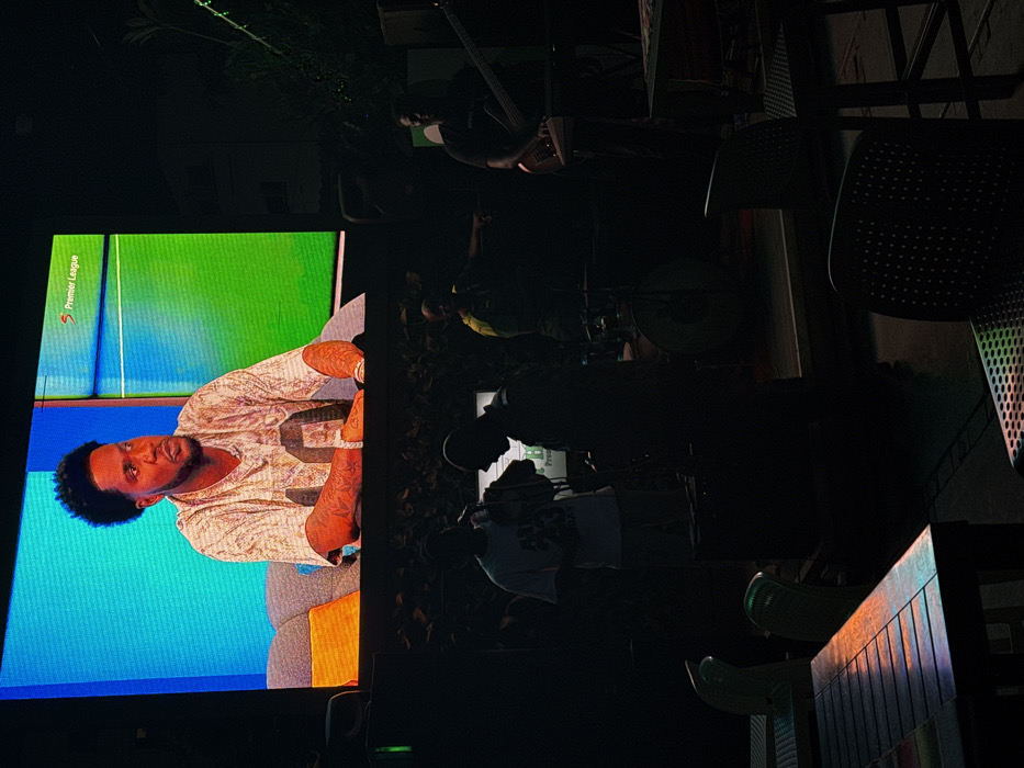 The image displays a dimly lit outdoor or semi-outdoor entertainment venue in Falomo, Nigeria. A large LED screen dominates the upper frame, showcasing a medium shot of an adult Black male with a dark, textured haircut and beard, wearing a light-colored, patterned short-sleeve shirt. Tattoos are visible on his forearms. He is depicted seated on a light gray sofa with a colorful cushion, against a background split into blue and green segments. The "Premier League" logo is visible in the upper right corner of the screen.
Below the screen, a group of at least five adult males are present in various states of silhouette, positioned on what appears to be a low stage or performance area. From left to right: one man in a light-colored t-shirt and pants holds an object, possibly a microphone or drink. Another man stands to his right, largely obscured by shadow. Further right, a third man is partially visible. A drummer and drum set are discernible in the center. To the far right, a man in a dark t-shirt is holding and appears to be playing a bass guitar or similar string instrument. Musical equipment, including speakers and drum components, is visible.
In the foreground, two empty tables and two empty chairs are visible. The leftmost table is wooden with a reflective surface displaying distinct red, orange, green, and blue light reflections. A light green chair is partially visible behind it. To the right, a dark, perforated chair also exhibits subtle colorful light reflections. The immediate background behind the performing individuals includes dark foliage or decorative greenery along a wall or structure. The overall scene indicates an evening or nighttime setting.