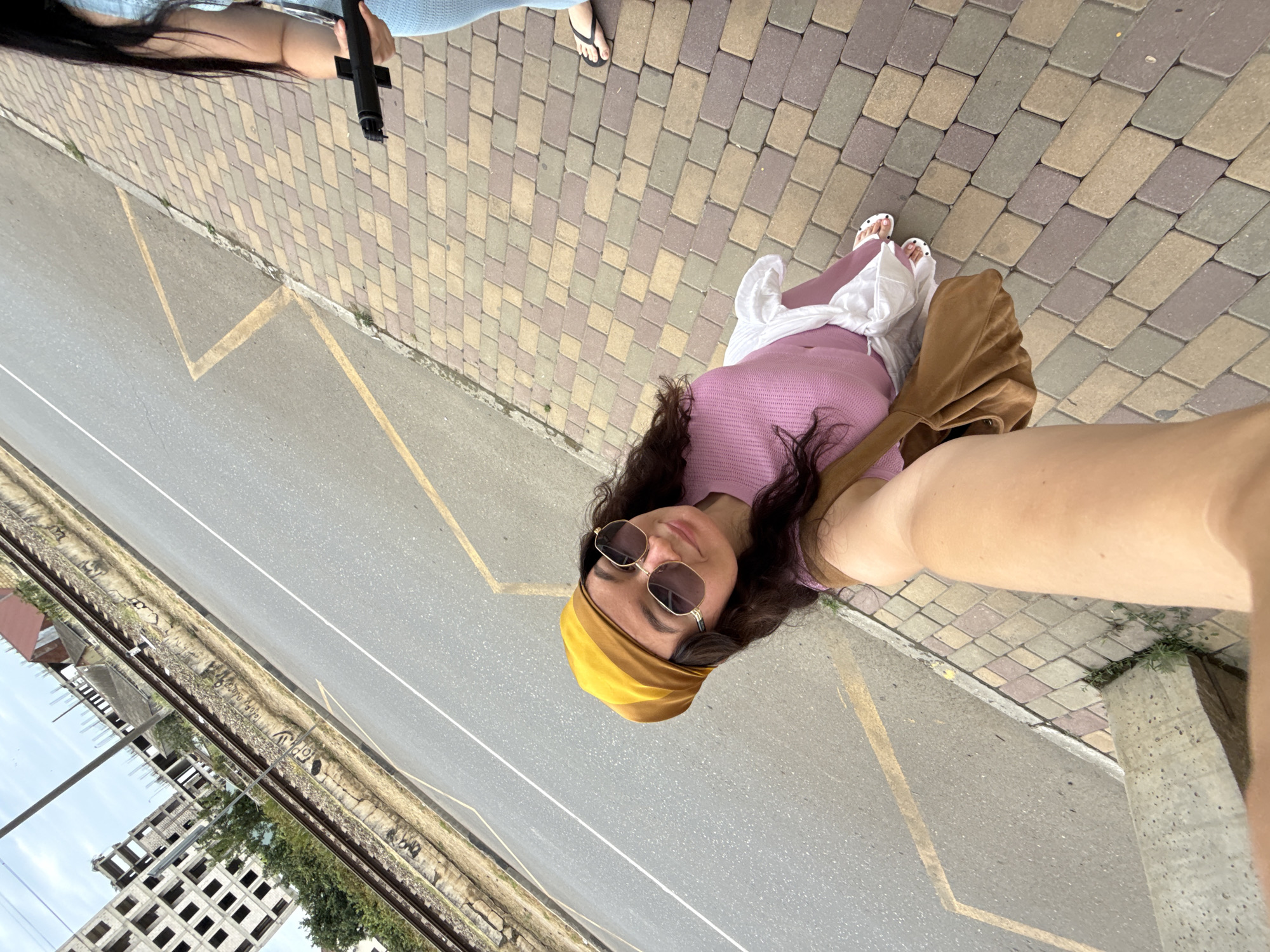 A first-person perspective, selfie-style image captures a young woman wearing a yellow headscarf, oversized sunglasses, and a pink top with a white skirt. She is holding a brown shoulder bag. The woman is standing on a paved walkway with multi-colored bricks, adjacent to a paved road with yellow markings. In the background, a set of train tracks runs parallel to the road, with a partially constructed building to the left and some houses to the right. Another person, partially visible on the right side of the frame, is wearing a blue outfit and black flip-flops, holding a black object. The scene appears to be an urban setting in Derbent, Russia, with a clear sky overhead.