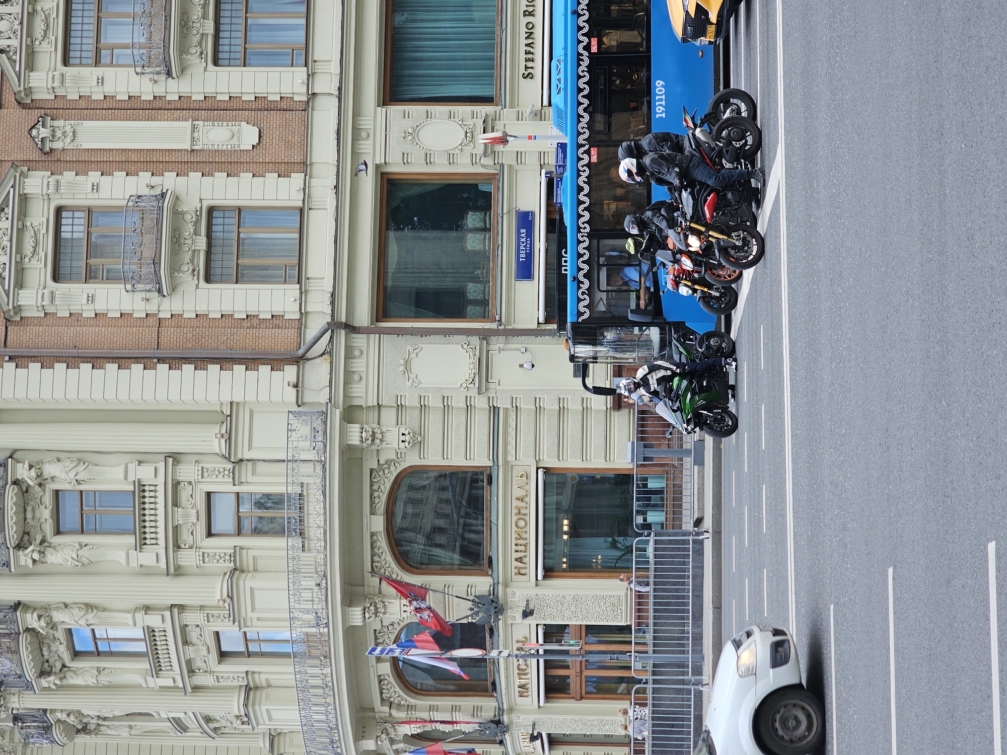 The image shows a street scene in Moscow, Russia, in front of the National Hotel. Several motorcycles with riders in protective gear are stopped at an intersection, with a blue bus behind them. A white car is also visible in the foreground. The National Hotel is a historic landmark in Moscow, known for its ornate architecture. The street is paved, and there are white lane markings. The sky is overcast.
