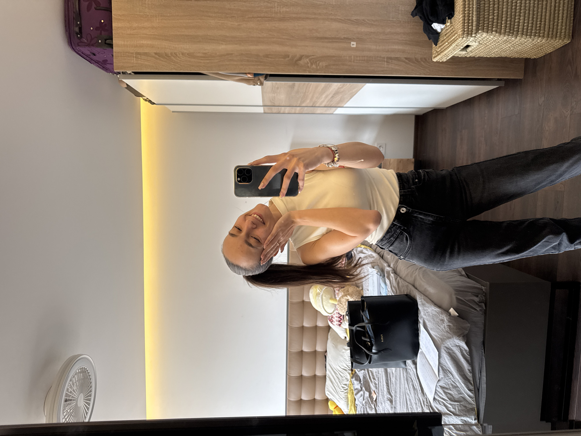 A young woman is taking a selfie in what appears to be a bedroom. She is wearing a white top and dark jeans. She is holding a smartphone in her right hand and smiling, while her left hand touches her face. Her dark hair is pulled back in a ponytail. The setting includes a bed with a gray comforter, a black tote bag on the bed, a closet with light wood and white doors, and a wicker basket on top of the closet. A purple suitcase is also visible on top of the closet. There is a wall fan and a strip of lighting on the wall. The location is likely an indoor setting. There are no details that indicate if this image was taken in Sofia, Bulgaria.