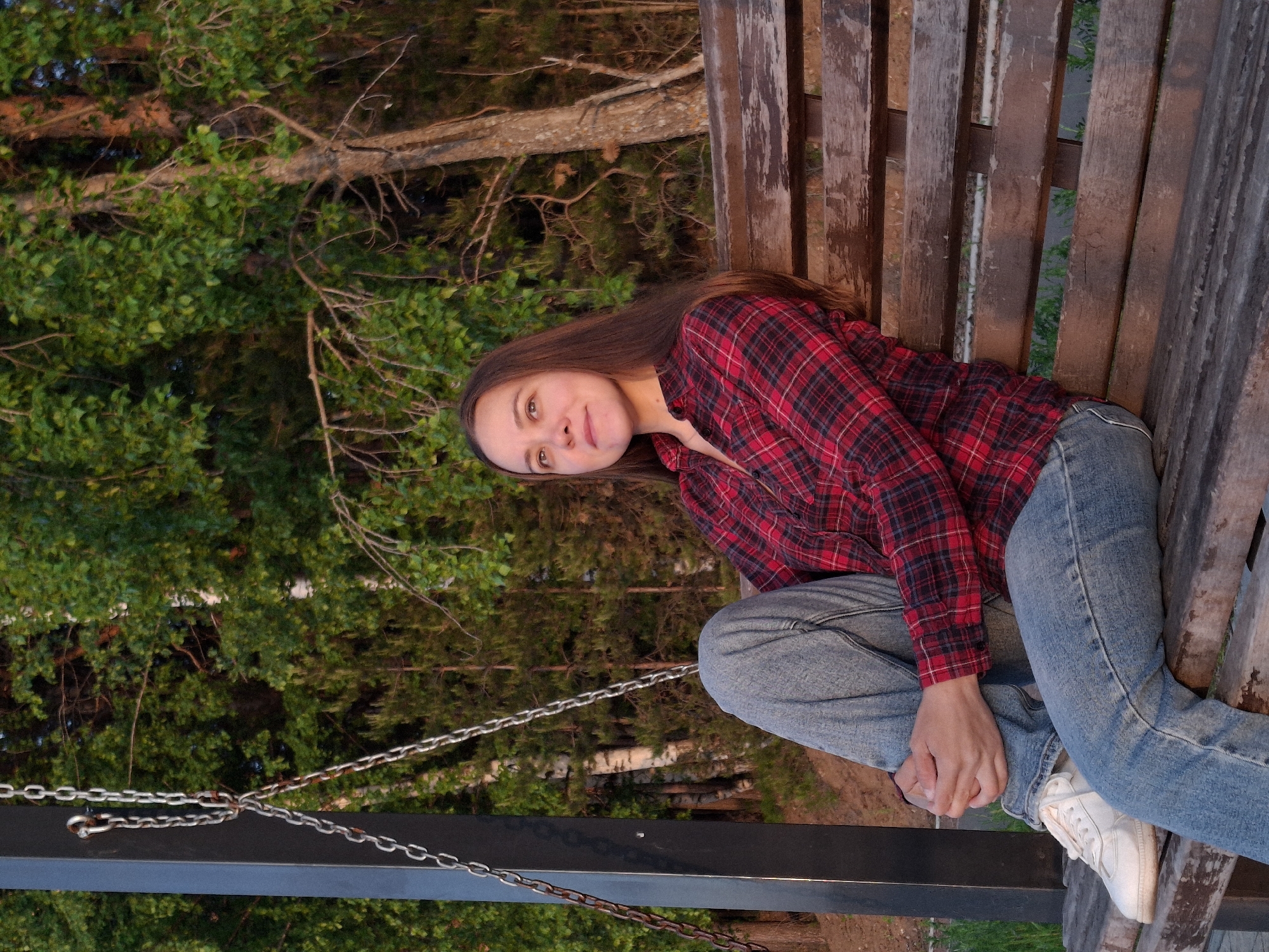 A young woman sits on a weathered wooden swing-bench in a park. She is wearing a red and black plaid shirt, blue jeans, and white sneakers. Her long brown hair falls over her shoulder. The swing is suspended by metal chains from a black frame. Behind the swing, there is lush green foliage of trees and branches. The scene appears to be in Chaikovsky, Russia.