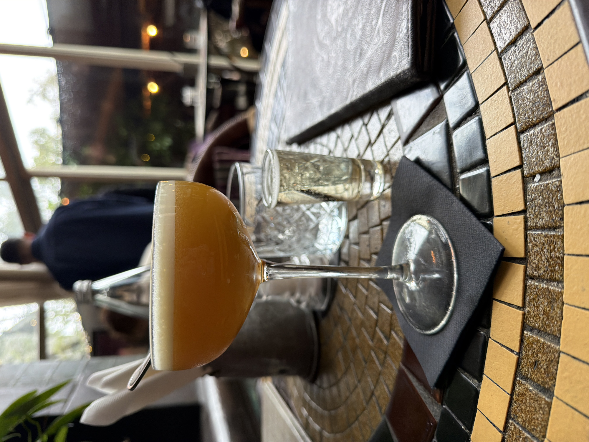 On a mosaic tile tabletop, a coupe glass filled with a yellow cocktail and topped with a thin layer of white foam rests upon a square black napkin. Adjacent to it is a small shot glass filled with a clear liquid. In the blurred background, a person in a blue shirt is visible. Through the window, trees and lights are seen. The setting seems to be indoors, possibly a bar or restaurant in Cape Town, South Africa. A bottle of water and some plants are also visible in the background.