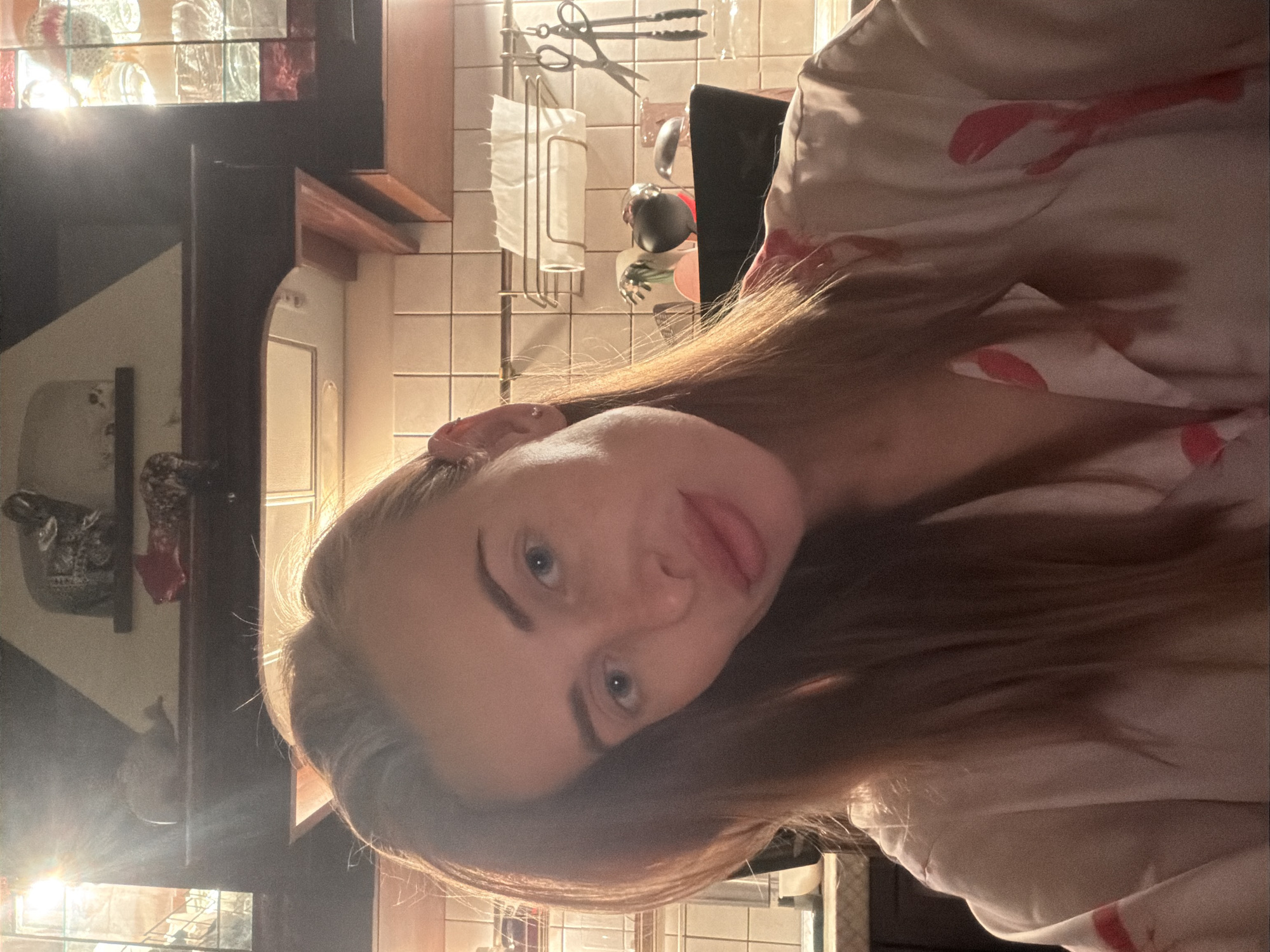 A close-up portrait of a person with fair skin, blue eyes, and light brown hair. The person is wearing a light-colored shirt with red patterns. The background appears to be a kitchen setting, with tiled walls, shelves holding utensils, and glassware.