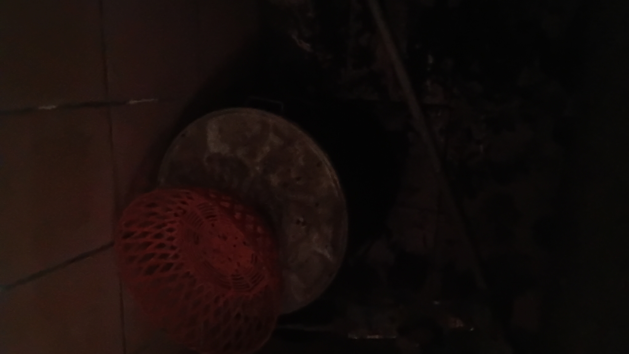 A red mesh basket rests on top of a metal pot. The pot appears to be empty and is sitting on a dark, uneven surface. The background is dark and appears to be an indoor environment with tiled walls. The overall lighting is dim. There are no people visible in the image. This scene is located in the city of Prabumulih, Indonesia.
