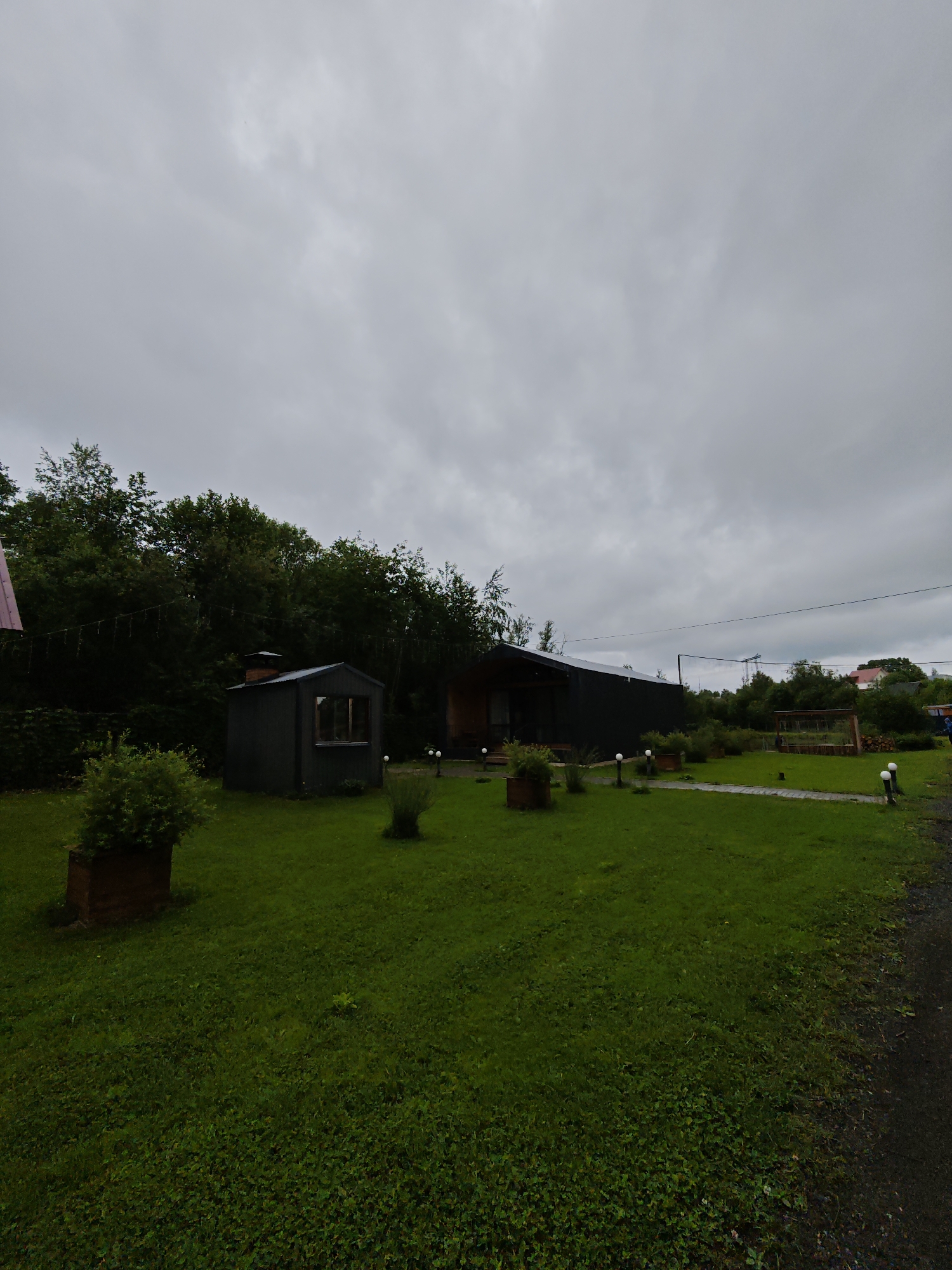 The image captures an outdoor scene in Malyi Bukor, Russia, under an overcast, light grey sky. The foreground and midground consist of a meticulously maintained, lush green lawn. A dark gravel or dirt path borders the right side of the lawn. A light-colored paved walkway extends from the lower left towards the mid-right, leading to a large building.
In the left-midground, a small, dark-colored shed-like structure is present, featuring a brown brick chimney on its left side and a rectangular window with a light frame on its front face. Reflections are visible on the windowpane. To the right and further back, a larger, modern building with dark-colored siding and a sloping roof is visible. Its front facade includes a covered porch area with a lighter-colored underside. Part of a reddish roof belonging to another structure is minimally visible on the far left.
Several spherical white outdoor lamps on short posts are strategically placed along the paved pathway and near the main building. Multiple dark-colored planters, each containing green shrubbery, are arranged on the lawn, along with a smaller standalone bush in the foreground. The background is characterized by dense green deciduous trees and bushes, suggesting a heavily vegetated or forested area. Utility lines are visible traversing the upper background against the sky. To the right of the main building, a wooden frame structure, possibly a pergola or garden feature, is discernable, alongside a stack of logs. No individuals are present in the scene.