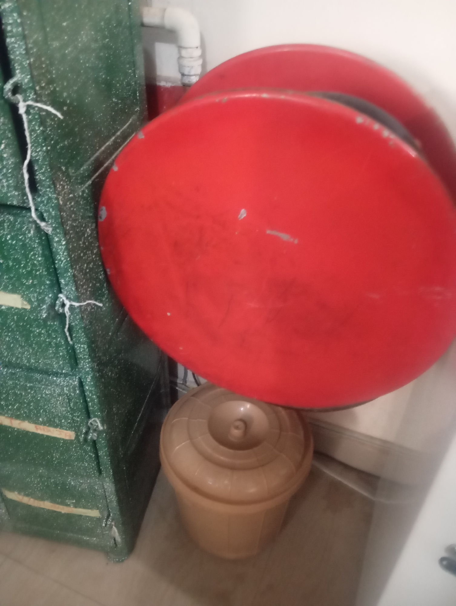 In an indoor environment, possibly a storage area or utility room, a large, round, red object dominates the foreground, obscuring much of the background. To the left, a green cabinet with a textured, speckled surface is partially visible, featuring yellow bands and metal latches. A thin white string is tied to the cabinet. Beneath the red object and to the right of the cabinet, a light brown, lidded plastic bucket is positioned. The overall impression is one of clutter or a utilitarian space. The location is not identifiable within the image.
