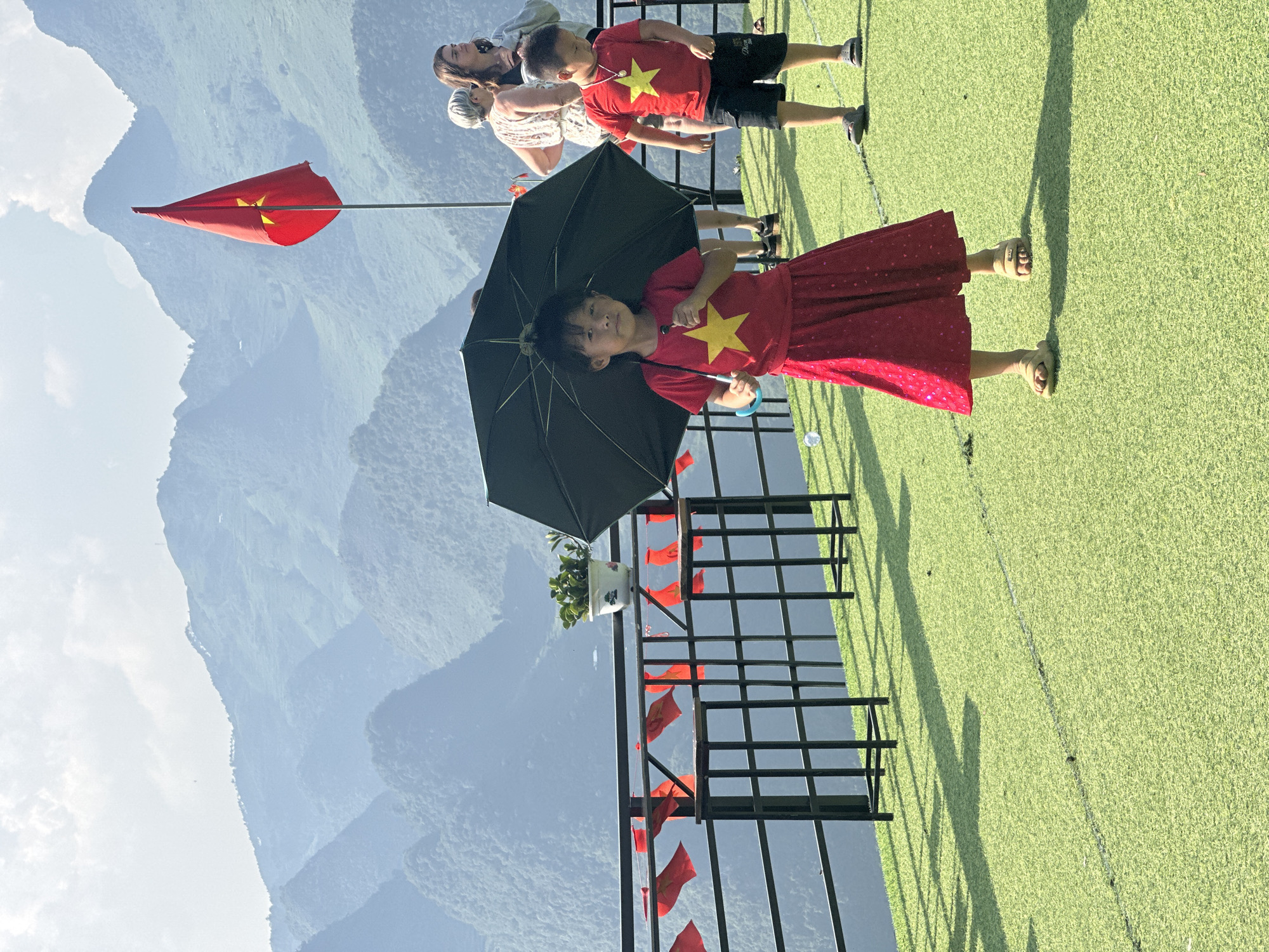The image depicts a scene in Đường Thượng Commune, Vietnam, with a child holding a black umbrella in the foreground, wearing a red dress with a yellow star, standing on green artificial turf near a black metal railing adorned with small Vietnamese flags. Other people, including a woman and a boy in a red shirt with a yellow star and black shorts, are standing behind the child. A large Vietnamese flag flies on a pole to the left. In the background, there are mountain ranges under a bright sky.