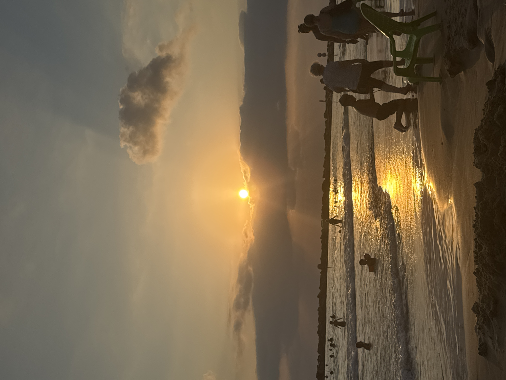 The image depicts a sunset scene at a beach in Netanya, Israel. Silhouettes of people are visible in the water and on the shore. The sun, partially obscured by clouds, casts a warm golden glow across the water and the wet sand. In the foreground, a green plastic chair sits on the beach. The water is calm, with gentle waves lapping at the shore. The overall atmosphere is serene and peaceful.
