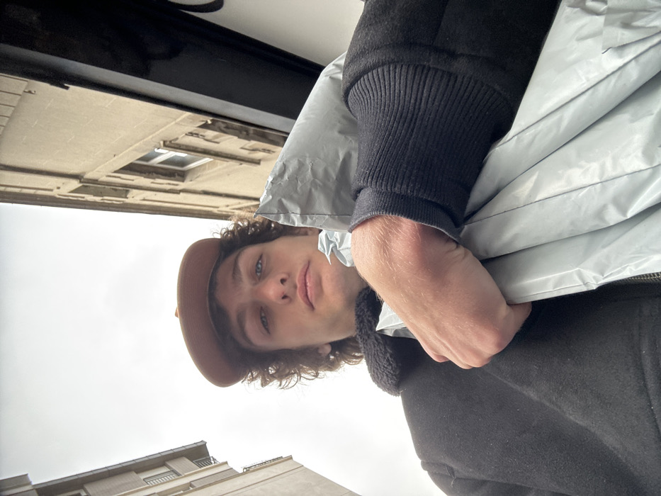 A low-angle, upward-facing image captures a young male individual in an urban environment in Joinville-le-Pont, France, under an overcast sky. The subject, centrally framed, has curly dark brown hair, blue eyes, and a neutral expression, gazing directly at the viewer. He wears a brown baseball cap and a dark-colored jacket with a black sherpa-style collar and ribbed cuffs. His left hand, positioned in the lower right foreground, firmly grips a crinkled, light grey, possibly plastic or synthetic, package or bag. The background shows two multi-story buildings flanking the open sky: a light-colored building with multiple windows on the left and a stone-textured building with similar windows and a dark vertical feature on the lower right on the right.