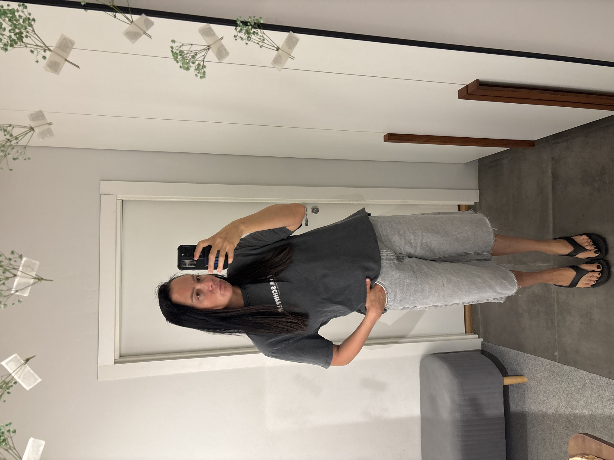 A woman is taking a mirror selfie indoors. The room has a white door with white trim, light grey concrete flooring, a light grey padded bench, and small flower arrangements taped to the mirror frame. The woman is wearing a dark grey t-shirt, light grey denim shorts, and black flip-flops. She is holding a black phone in her right hand and posing with her left hand on her hip. The reflections on the wall show two narrow wooden beams. The location details (city of Chaikovsky, Russia) cannot be verified from the visual data.