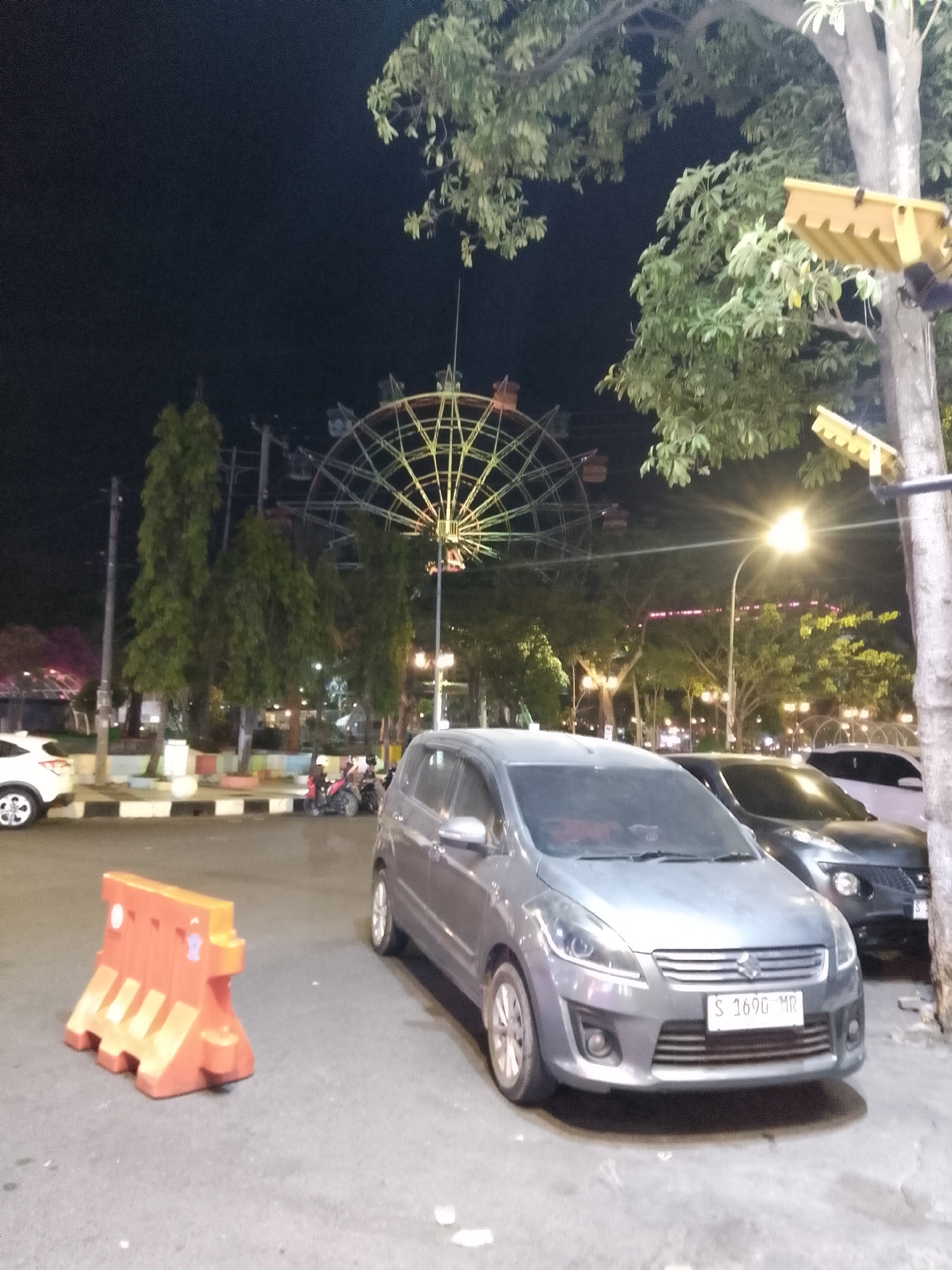 At night, a grey car is parked on a street, facing the viewer. The car has a license plate that reads "S 1690 MR". Behind it, another car is parked, facing away from the viewer. In the background, a Ferris wheel is illuminated with colorful lights. The scene appears to be in Lamongan, Indonesia, likely at a night market or fair. An orange plastic barrier is on the left side of the image, and trees and streetlights are visible throughout the scene.