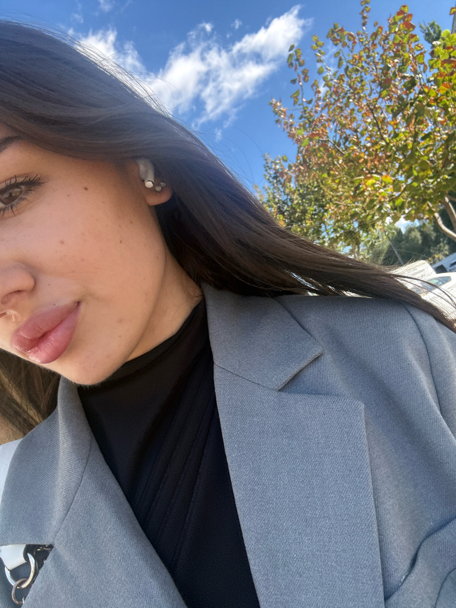The image is a close-up, outdoor, selfie-style shot of a young woman. She has long, dark brown hair, brown eyes, and light skin with some freckles. She is wearing a gray blazer over a black turtleneck top. She has on an earbud in her right ear and a small stud earring. Her lips are slightly parted and appear to have a gloss or balm on them. The background shows a blue sky with scattered white clouds and the leaves of a tree with some autumnal colors. The lower right portion of the image shows the lapel of her blazer. The lighting suggests it is daytime. There is no identifiable urban infrastructure or landmarks visible that would place the location in Chișinău, Moldova.