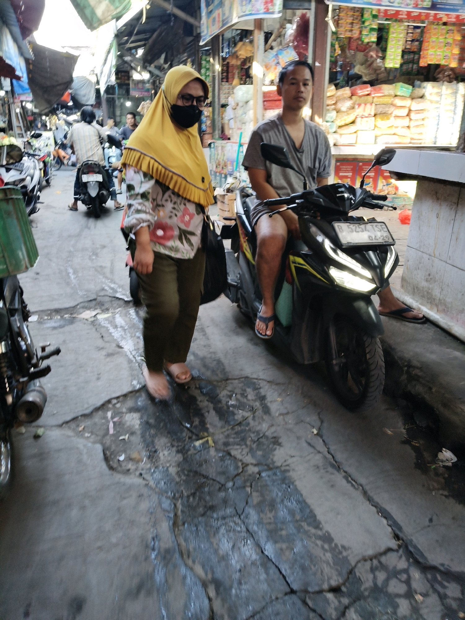 In the city of Setiamekar, Indonesia, an outdoor market scene unfolds with a focus on two individuals and their motorcycles. The foreground is dominated by a black scooter with a license plate reading "B 5254 TSE 04-29." A man, seated on the scooter, wears a grey t-shirt and dark shorts, with his legs exposed. He is looking towards the camera with a neutral expression.
To the left of the scooter, a woman is partially visible, walking away from the camera. She is wearing a yellow hijab, glasses, and a black face mask, obscuring her facial features. Her attire includes a floral-patterned shirt and olive-colored pants. Her feet are bare, with one sandal visible.
Behind these two individuals, the market environment is evident. Stalls with various goods, including packaged food items, are visible in the background. Other motorcycles and people are present, suggesting a bustling marketplace atmosphere. The ground is paved and appears somewhat wet and cracked in places. The lighting suggests it is daytime, possibly late afternoon or early morning.